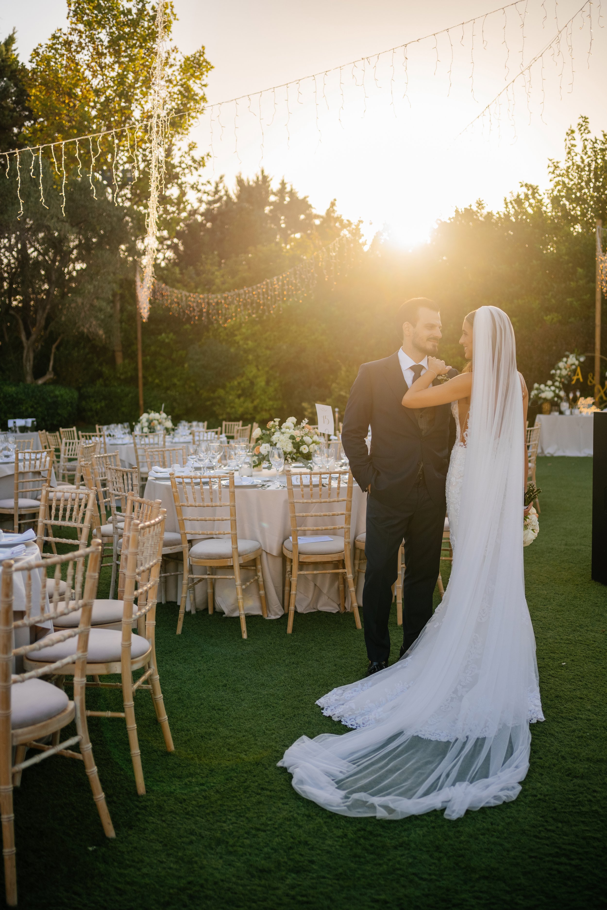 A bride and groom standing together at an outdoor wedding reception during sunset. The bride is wearing a white wedding gown with a long train and veil, and the groom is in a dark suit. There are decorated tables and chairs with white tablecloths and