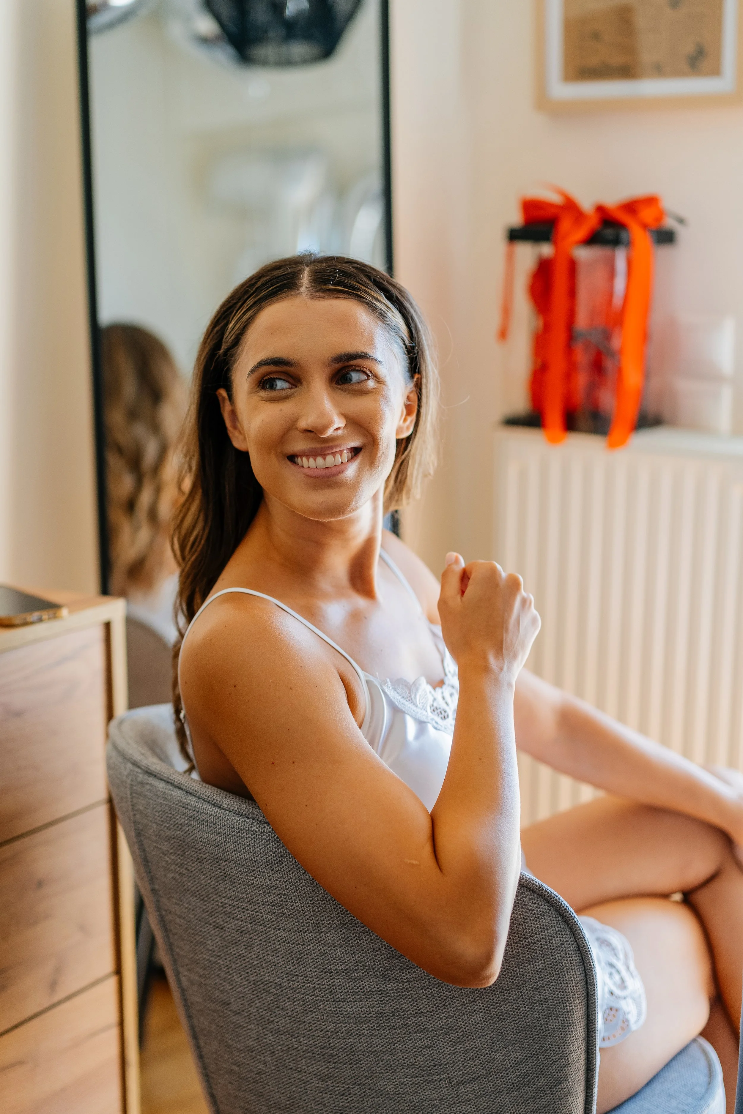 A smiling woman with brown hair and blue eyes sitting in a gray chair, wearing a white lace camisole, in a room with a wooden floor, a mirror, and a shelf with gifts tied with orange ribbons.
