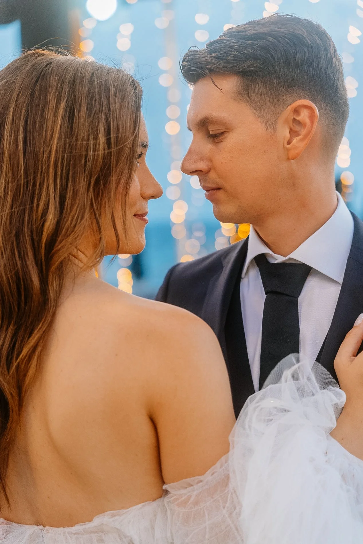 A couple dancing closely at a wedding reception, with the woman wearing a strapless white dress and the man in a black tuxedo, against a background of blurred string lights.