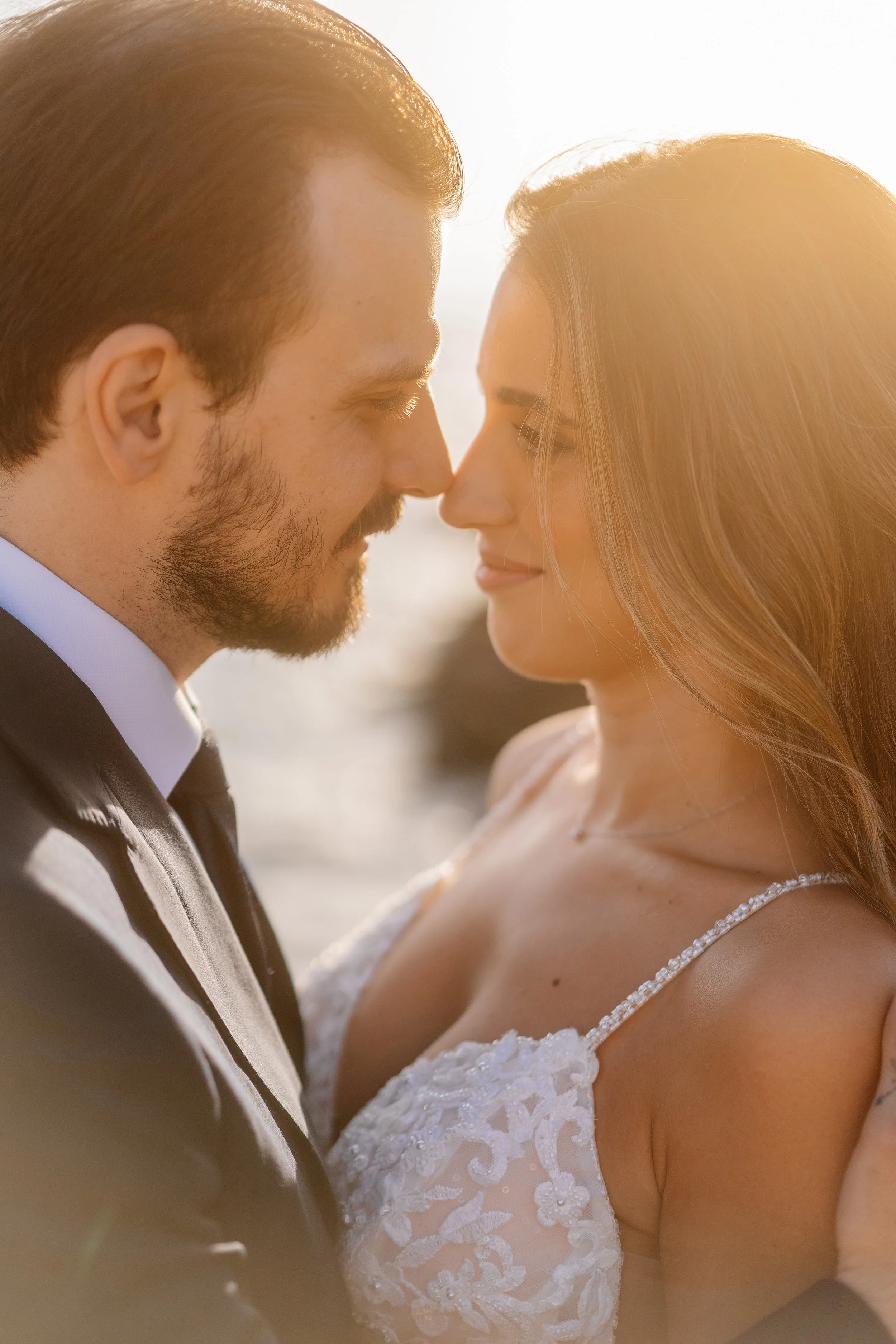 A close-up of a couple on their wedding day, touching foreheads, with soft sunlight backlighting them, both smiling gently.