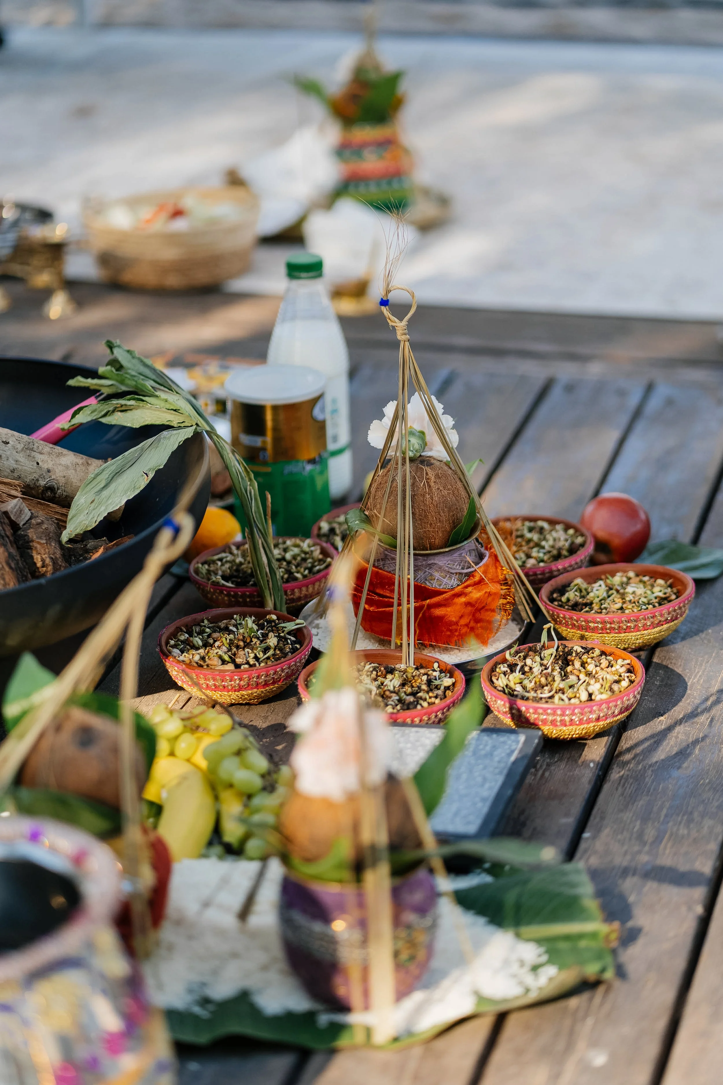 A traditional Hindu or Buddhist offering setup on a wooden surface, with bowls filled with grains, flowers, and a coconut decorated with leaves and a cloth, likely for a ritual or prayer.
