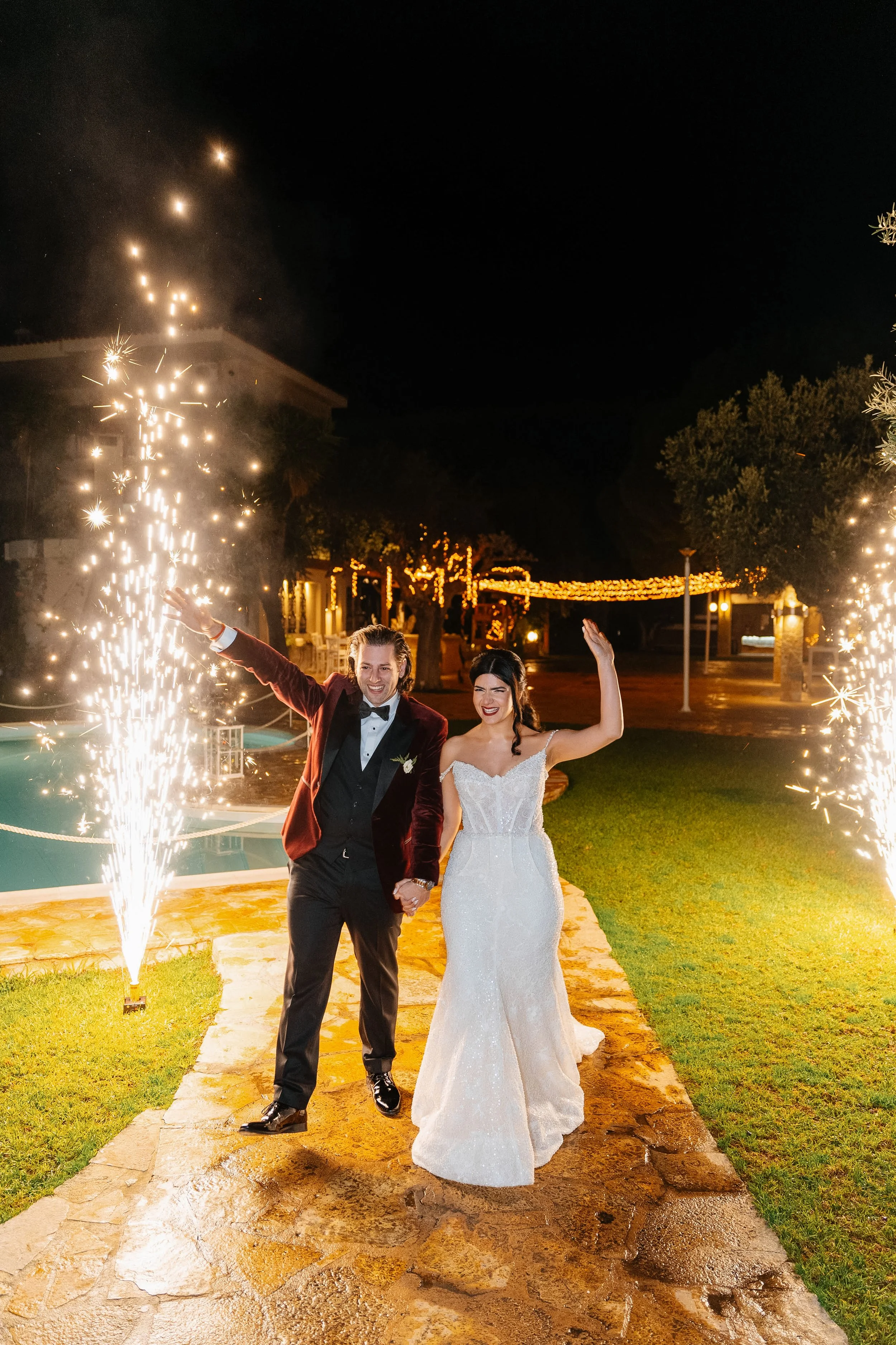 A newlywed couple walking hand-in-hand outdoors at night, celebrating with fireworks. The groom is wearing a dark suit with a bow tie, and the bride is in a white strapless wedding gown, both smiling.