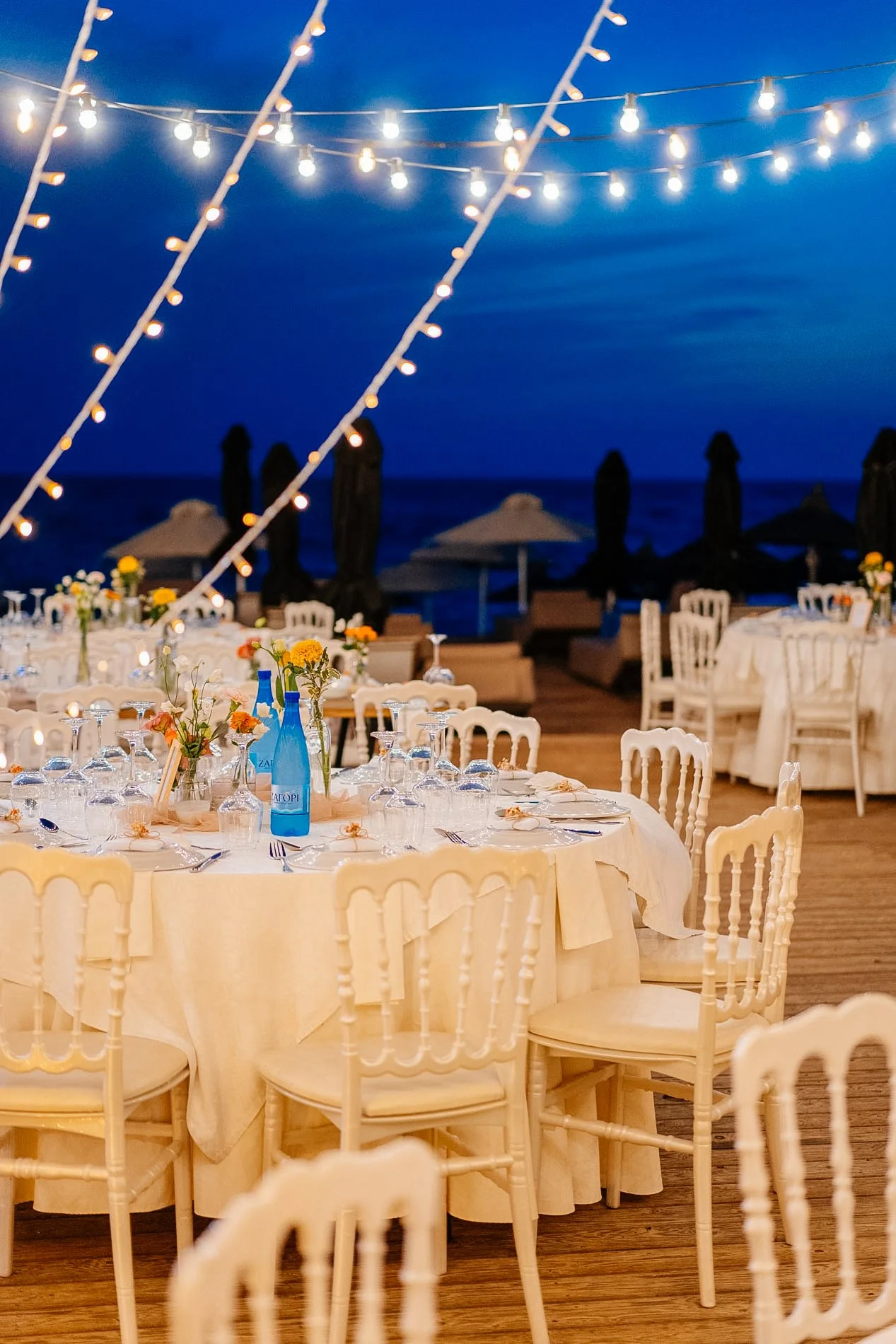 Decorated outdoor dining area at night with string lights, floral centerpieces, and ocean in the background.