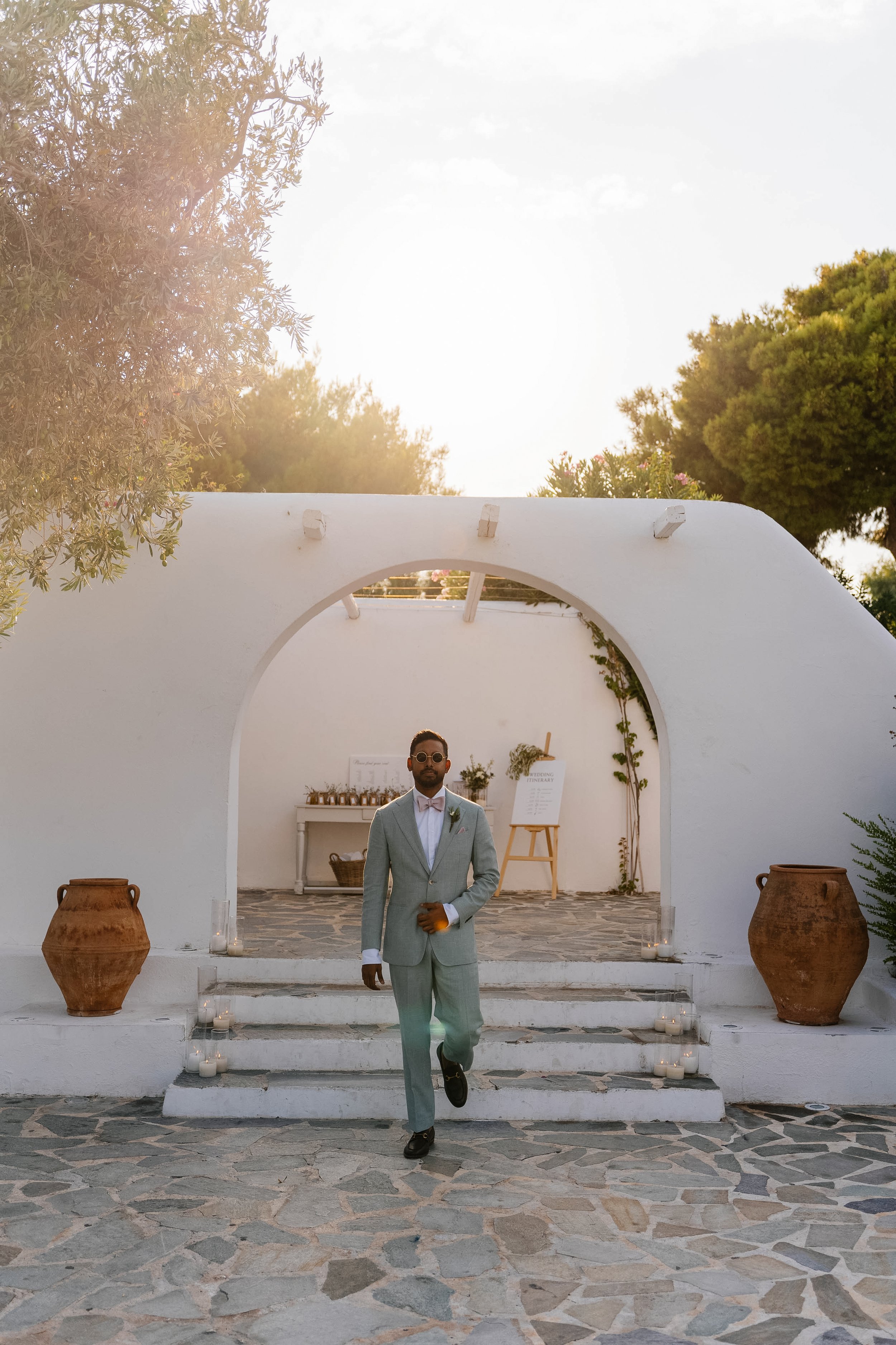 A man in a light gray suit with a pink bowtie and sunglasses walking down steps decorated with candles. The background features a white building with an archway, potted plants, and trees, under a bright sky with some clouds.