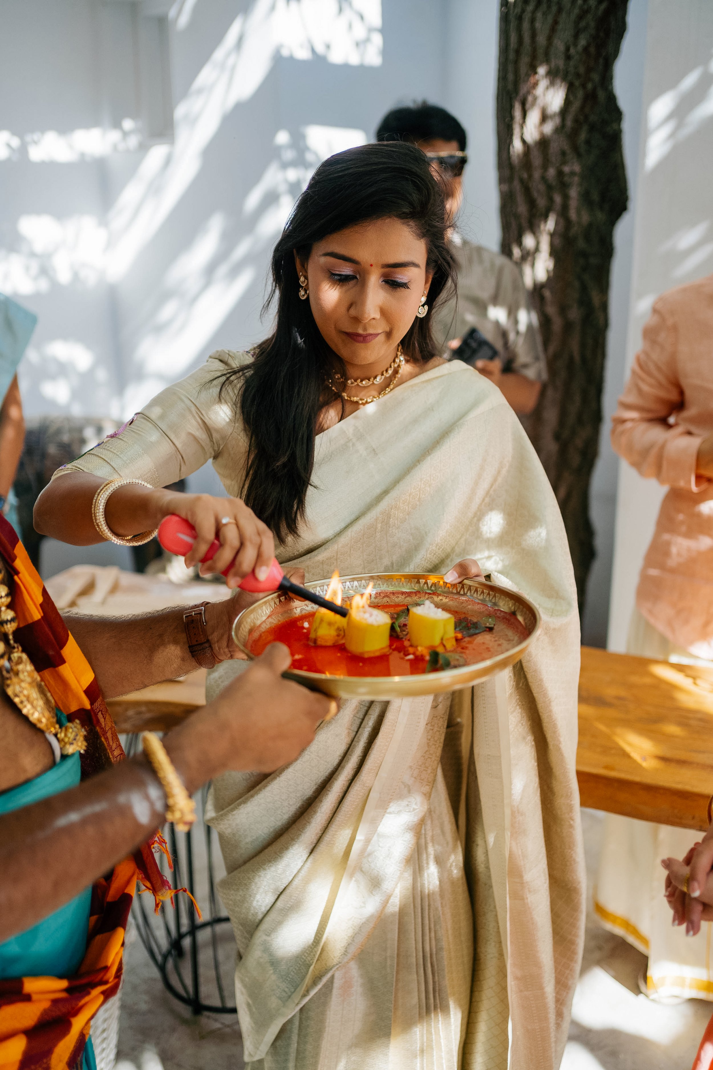 A woman dressed in traditional Indian attire is participating in a ceremony where a person is pouring a liquid onto a small flame on a round tray, with candles and flowers. She is standing outdoors under sunlight and shadows of a tree are visible.