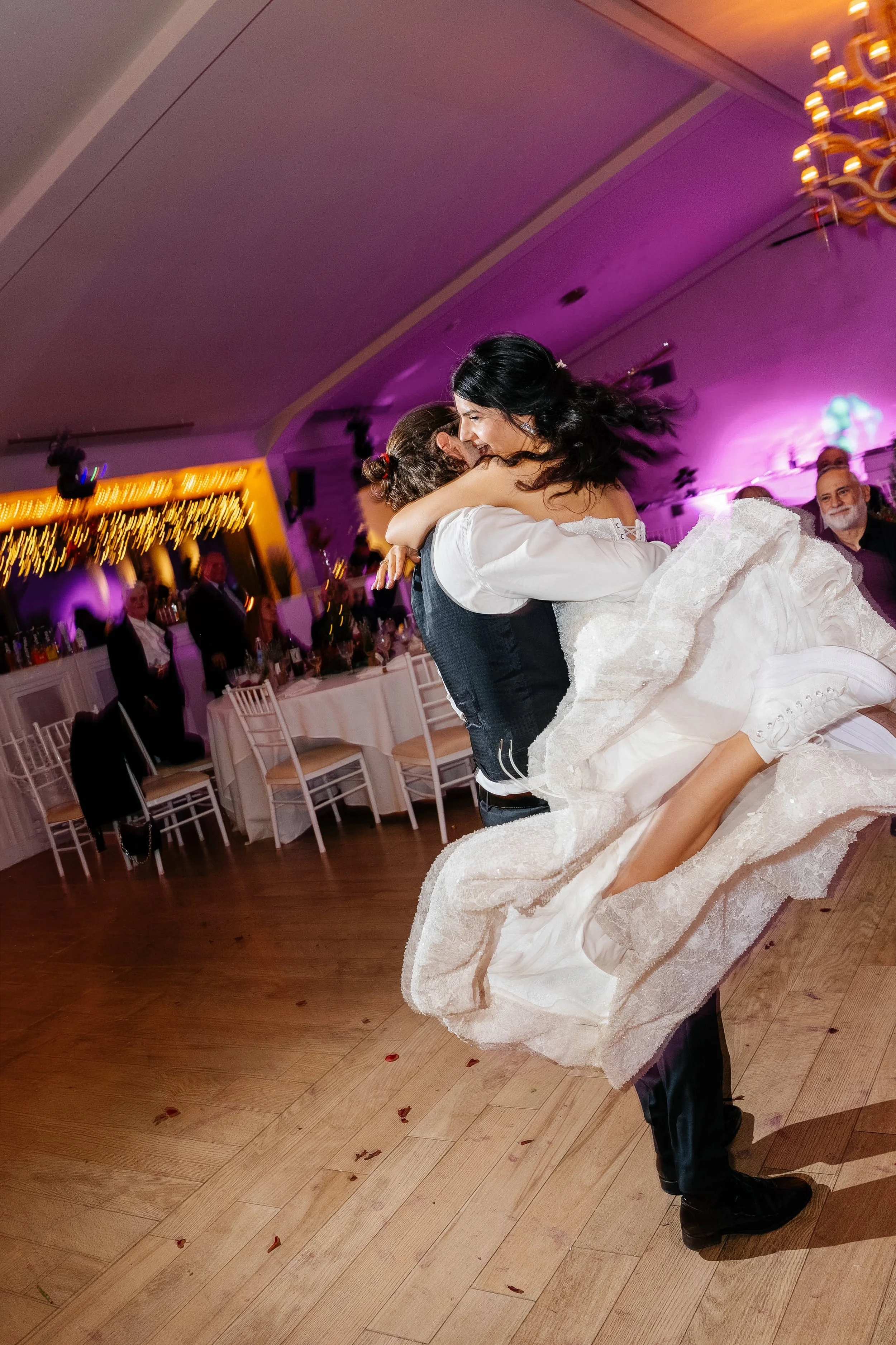 A man in formal attire dancing with a woman in a wedding dress at a wedding reception. The woman is being lifted and they are smiling. The background shows guests, tables, chairs, and colorful lighting.