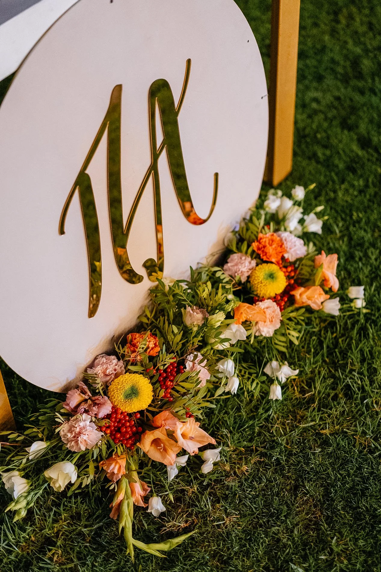 Close-up of a white circular sign with the initials AXY in gold, surrounded by colorful flowers and greenery on grass.
