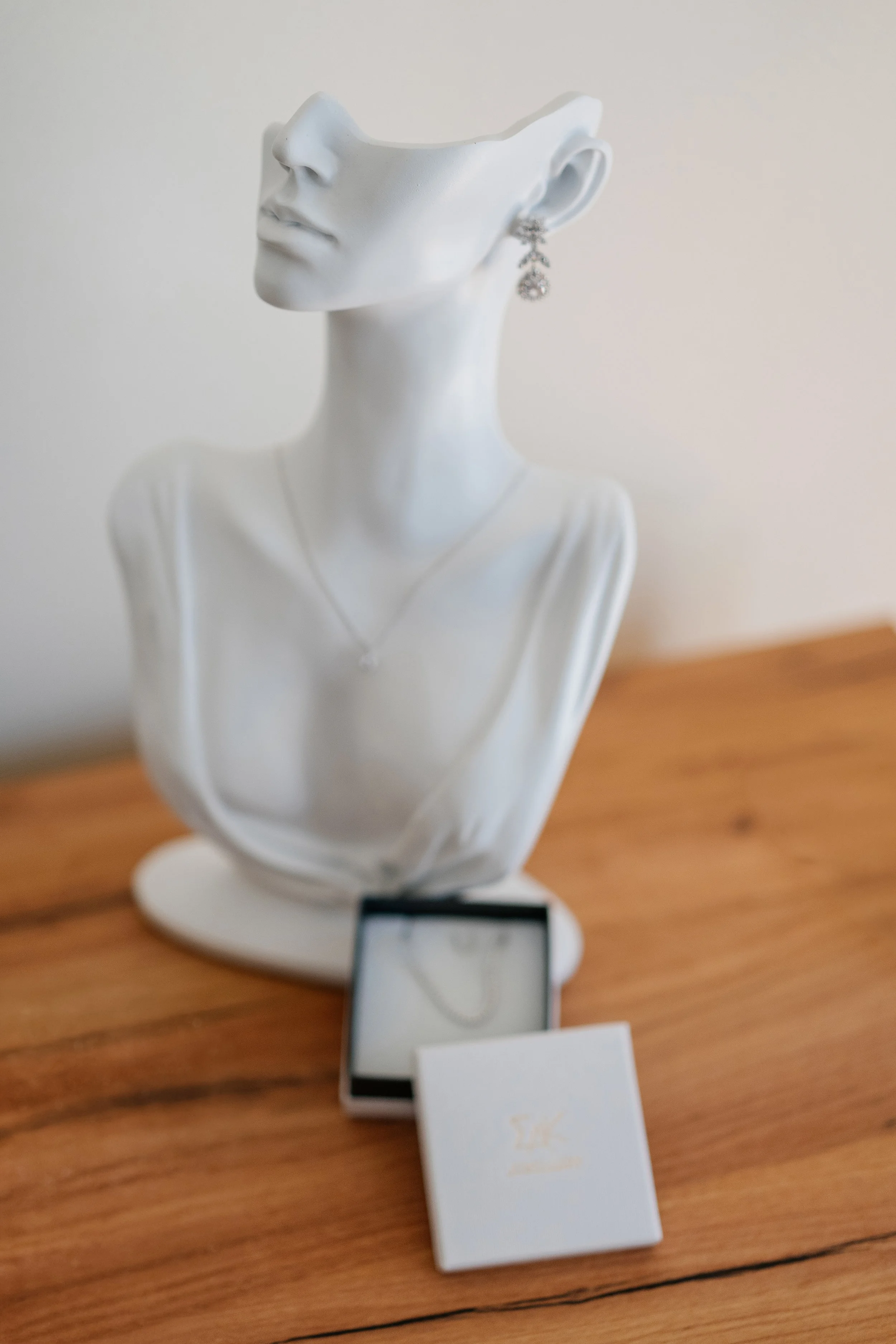 A white mannequin bust with an earring and a necklace, displayed on a wooden surface, with a small smartphone and a jewelry box in front.