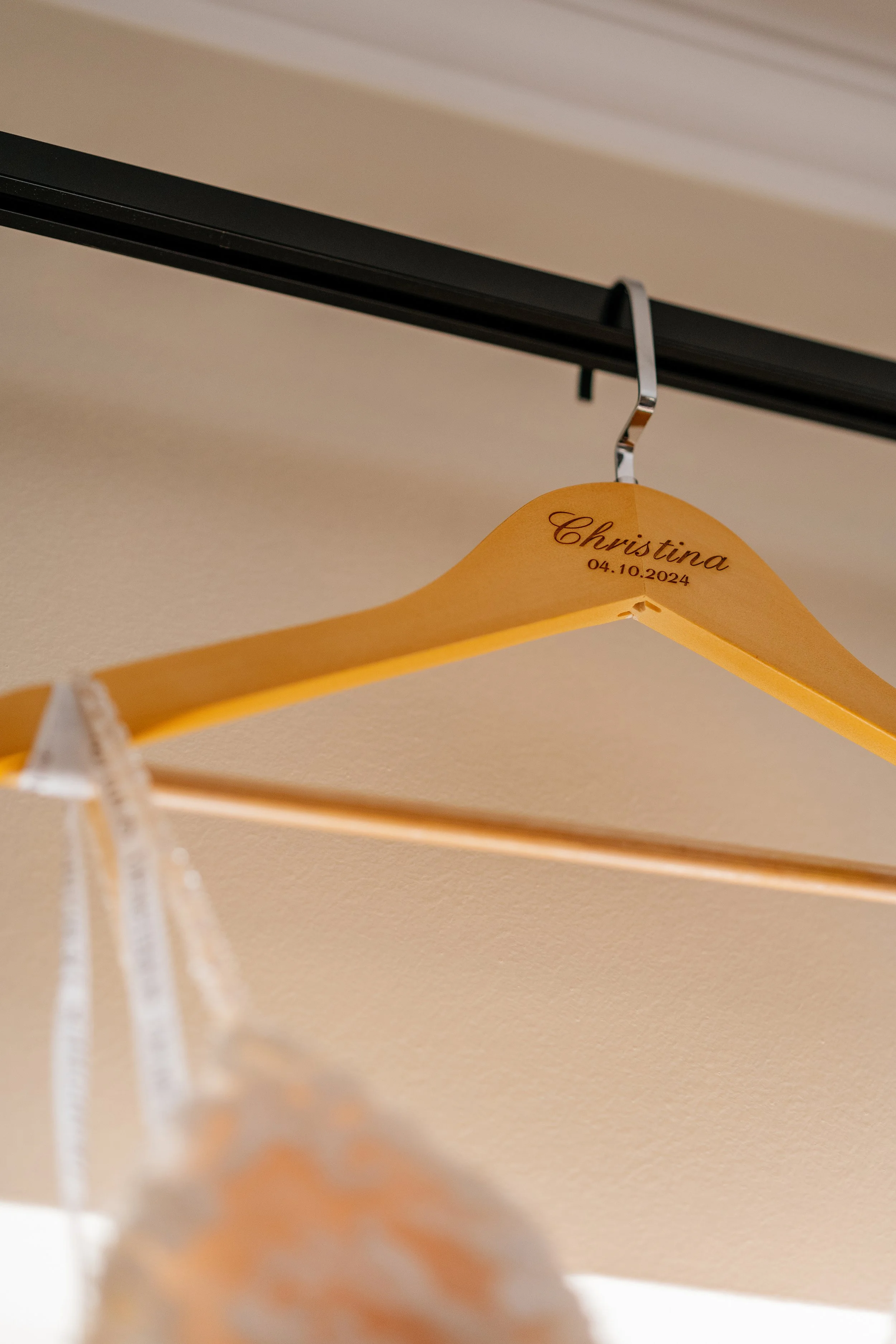 A wooden hanger with the name 'Christina' and the date '04.10.2024' printed on it, hanging from a metal hook on a black horizontal rod.