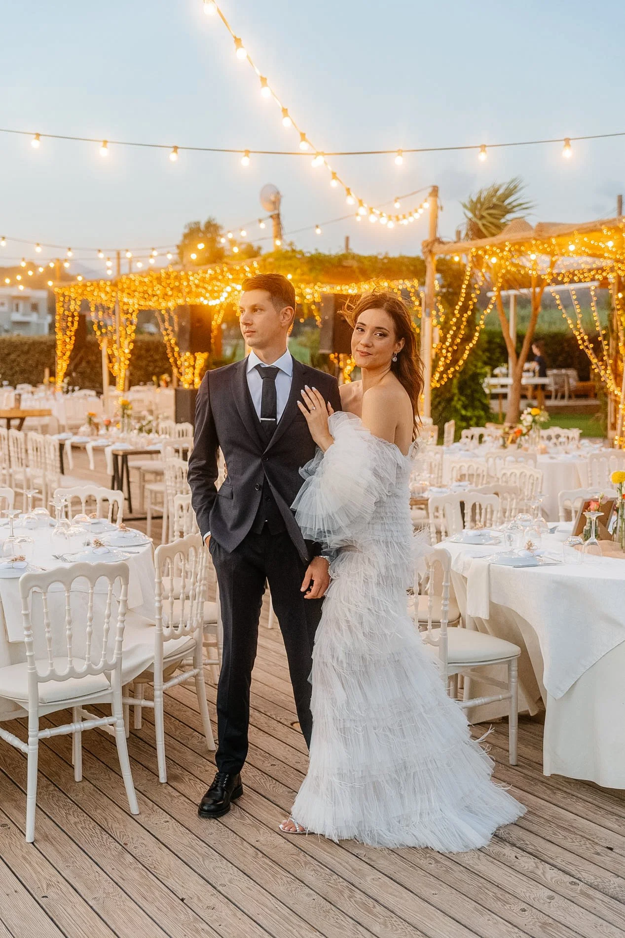 A bride and groom at a wedding reception outdoors during evening, with string lights and decorated tables in the background.