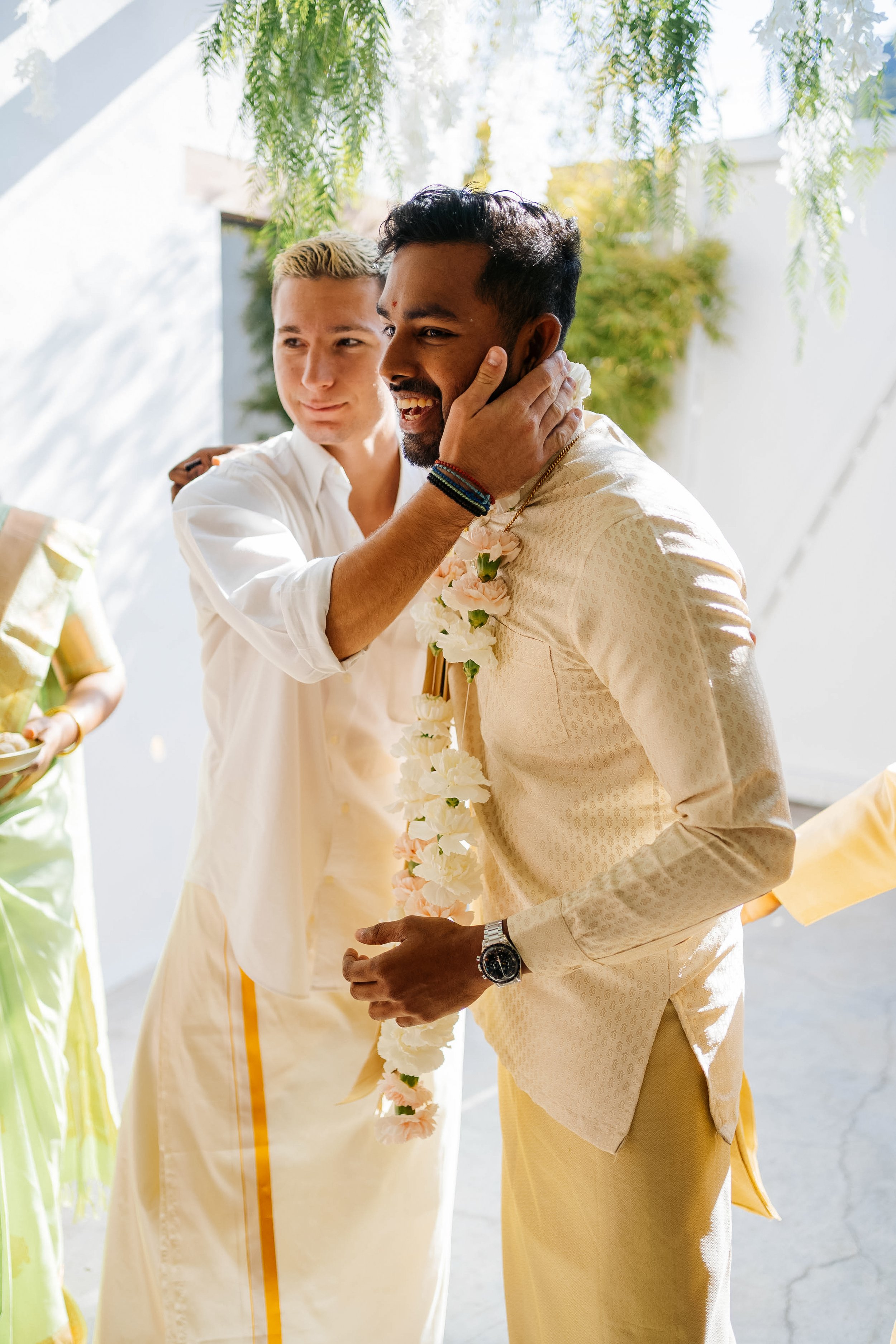 Two men are dressed in traditional Indian wedding attire, with one placing a garland around the other's neck. They are smiling and touching each other's faces, in a festive outdoor setting with greenery and bright natural light.