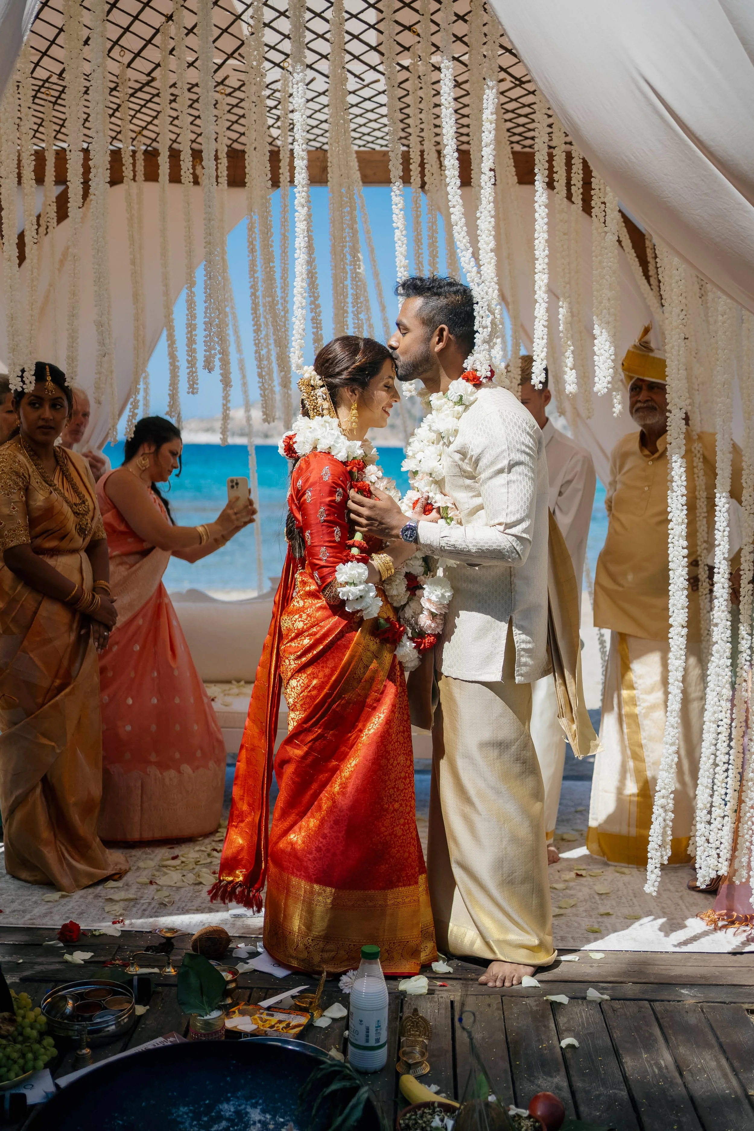 A couple in traditional Indian wedding attire shares a Kiss during a beach wedding ceremony, under a decorated canopy with flower garlands, surrounded by their guests.