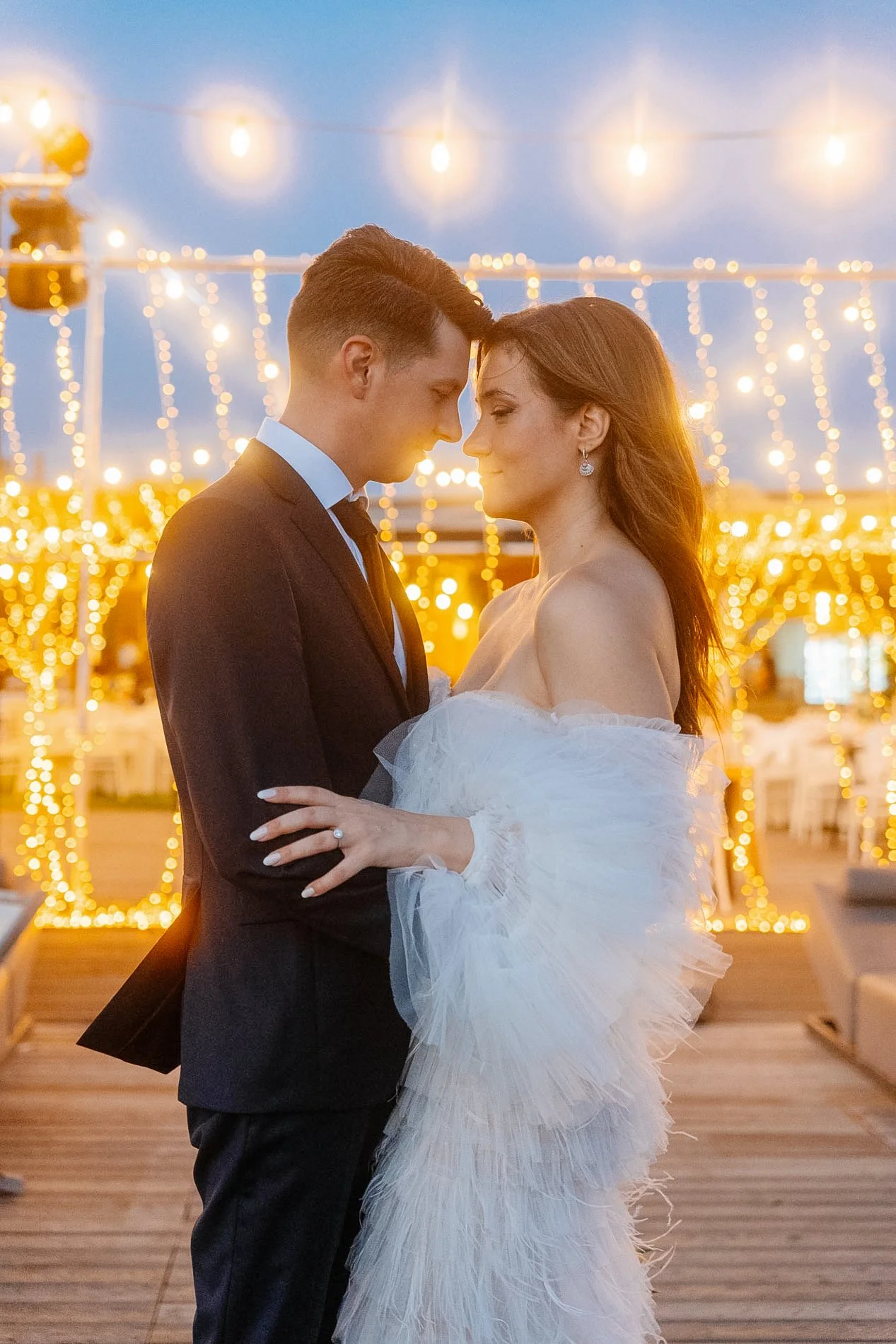 A bride and groom standing close with foreheads touching during their wedding reception, illuminated by string lights in the background.