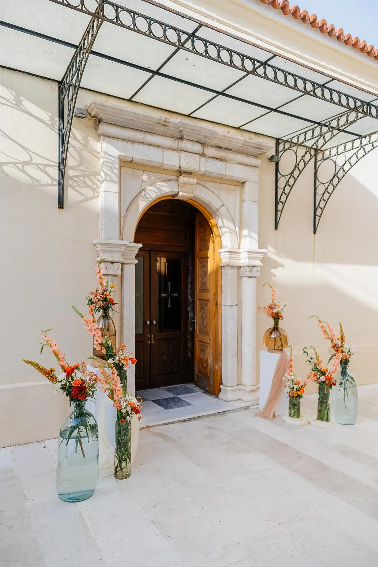Entrance decorated with flower arrangements in glass vases, a wooden door with an arched stone frame, and a porch with decorative metal supports along the roof