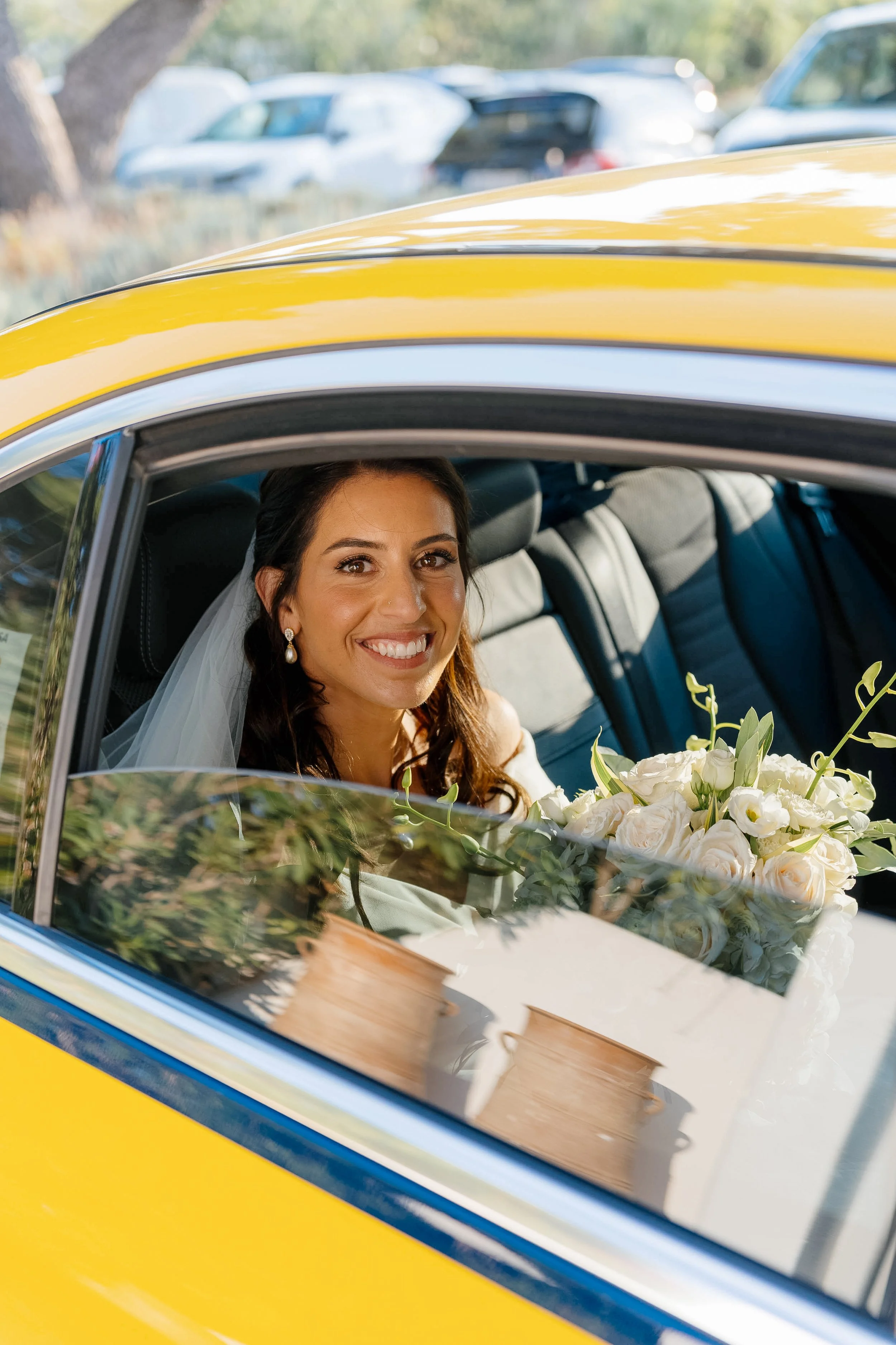 A smiling bride with dark hair and pearl earrings sitting in a yellow taxi, holding a bouquet of white flowers, with blurry cars and trees in the background.