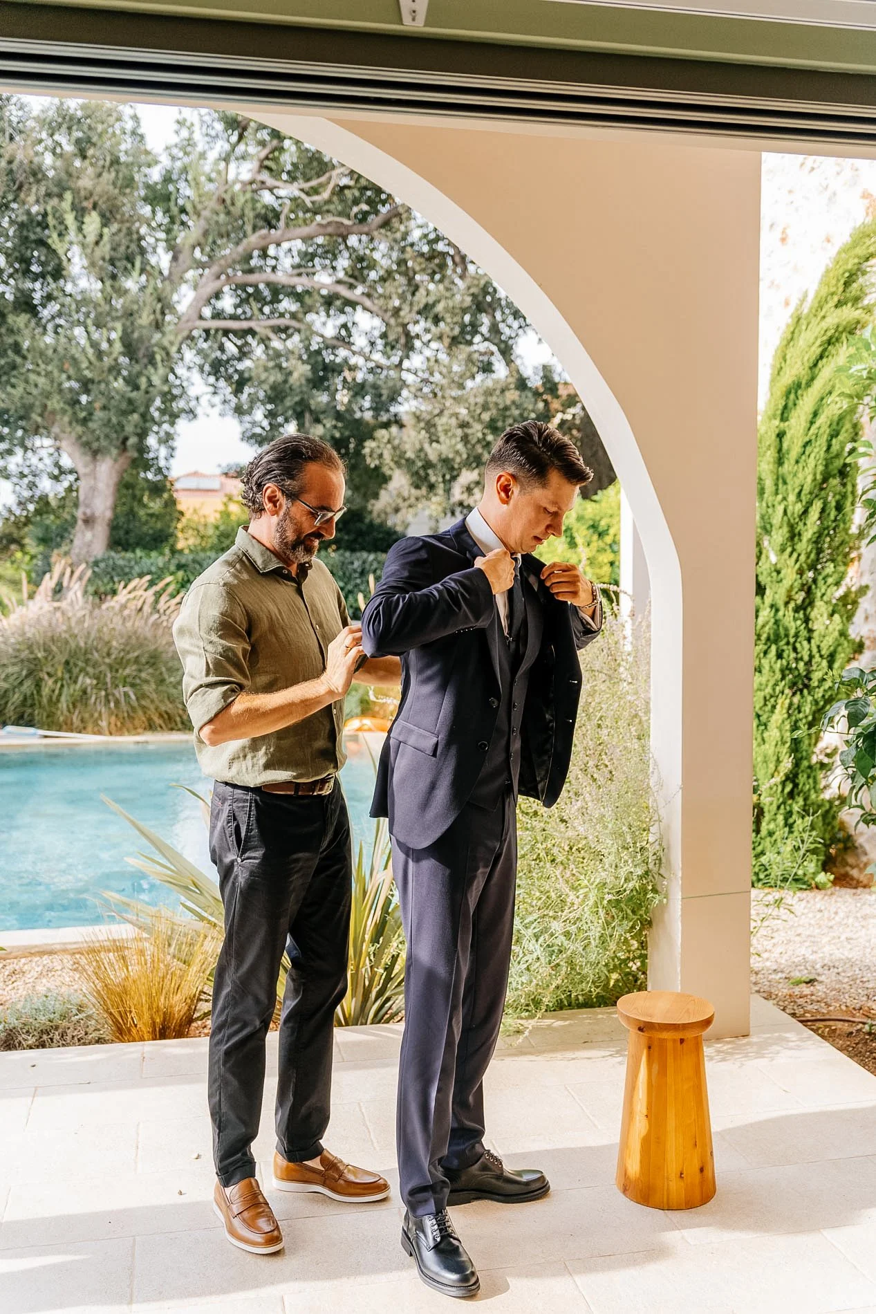 A man in a suit is adjusting his jacket while another man, dressed casually, looks on, outside near a pool and greenery.