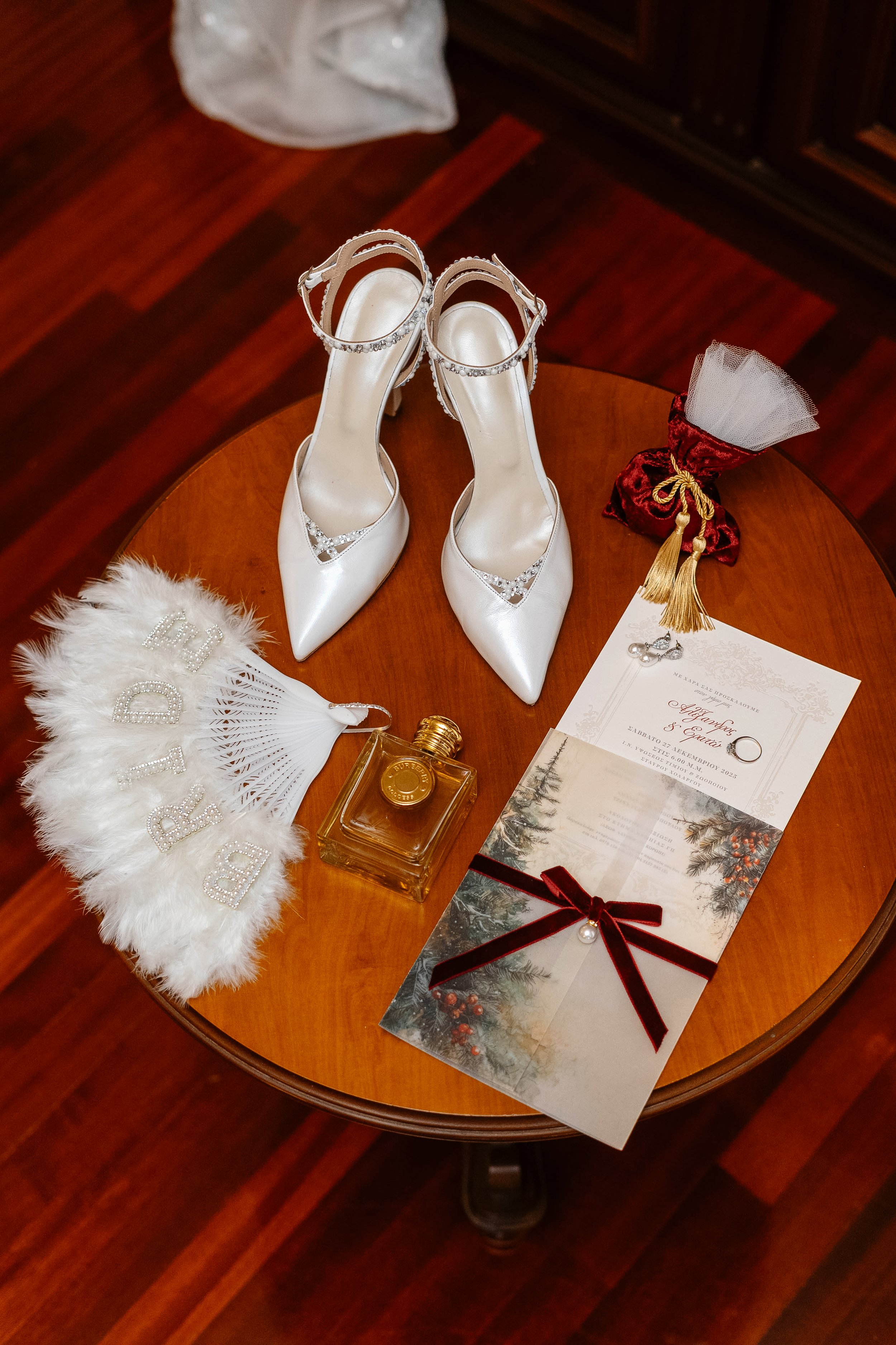 Array of wedding accessories on a round wooden table including white high heels decorated with rhinestones, a white feathered fan with 'Love' spelled in rhinestones, a small red velvet gift bag with white tulle, a pair of pearl earrings, a perfume bo