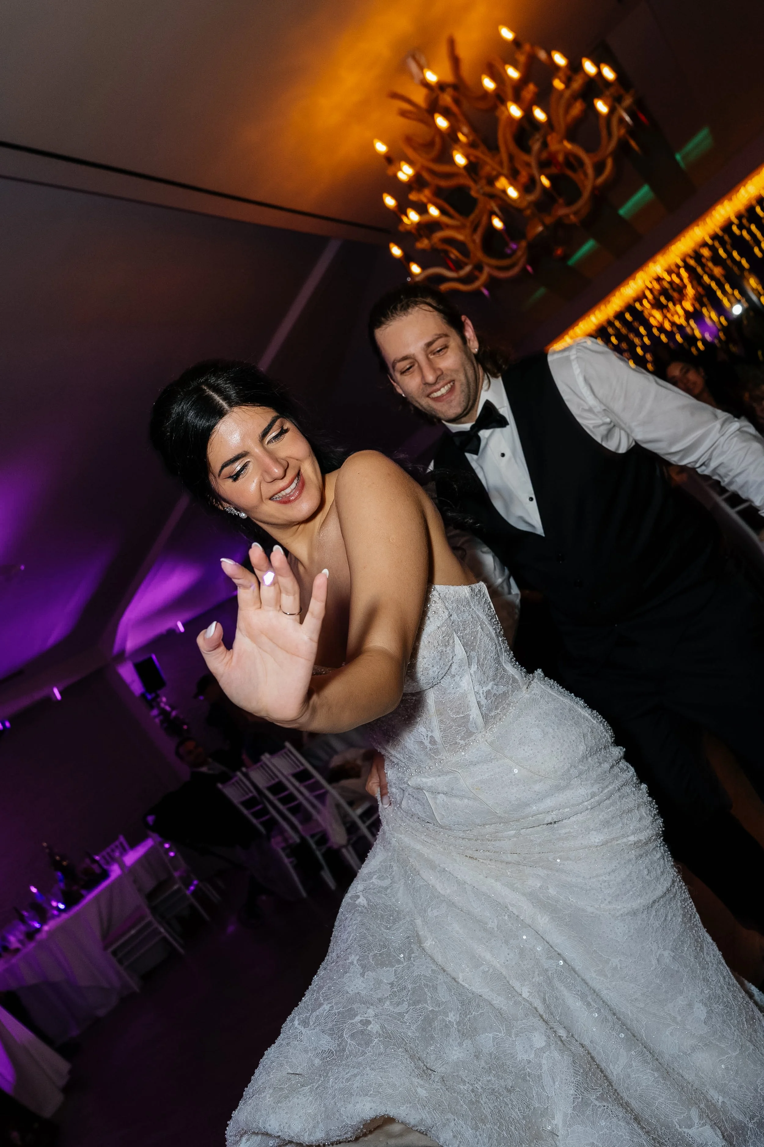 A bride and groom dancing at their wedding reception in a dimly lit ballroom with a large chandelier and purple lighting.
