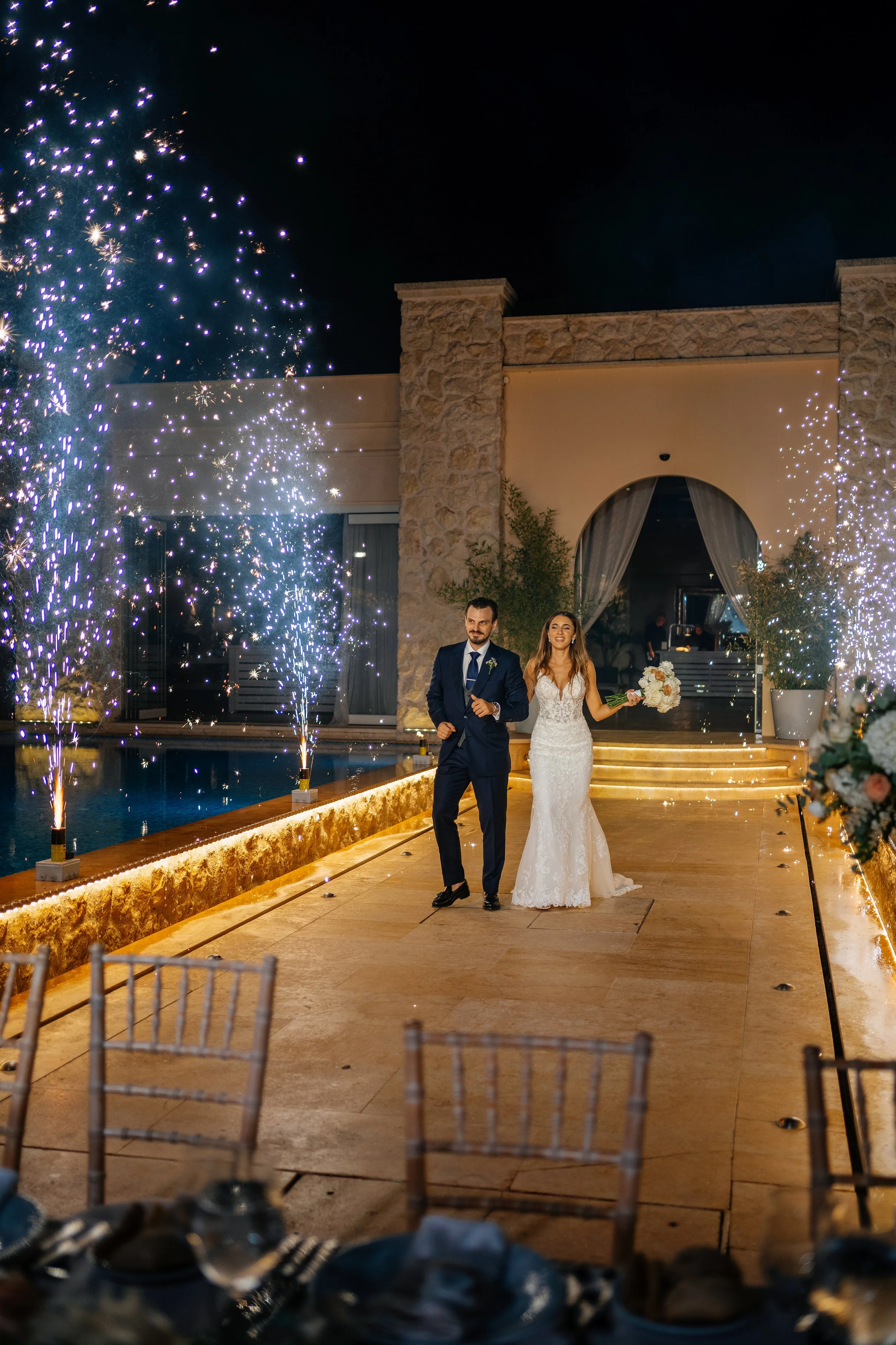 A newlywed couple walking down a decorated outdoor aisle at night, with fireworks in the background, celebrating their wedding.