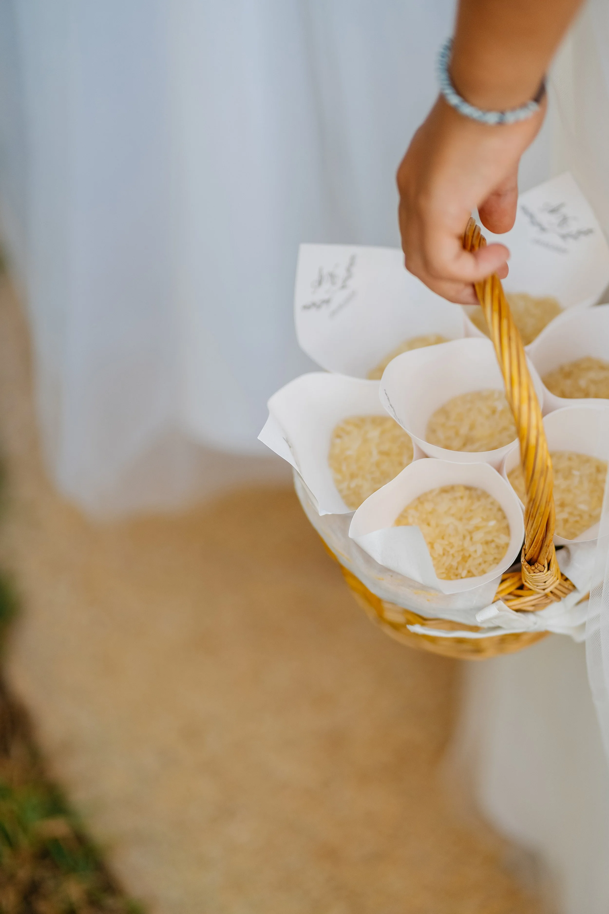 A hand with a bracelet selecting cookies from a basket lined with white paper cups, on a beige surface.