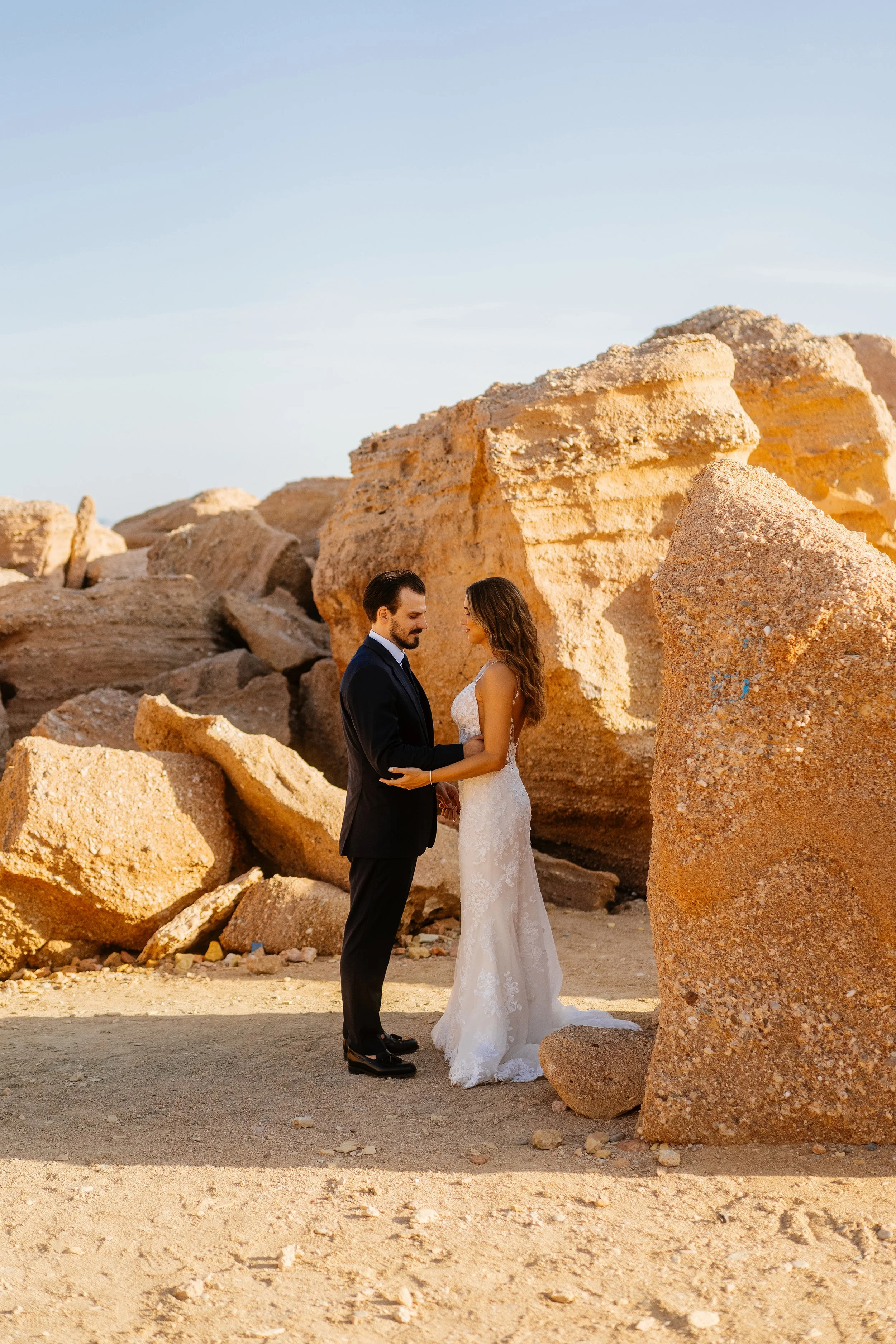 A bride and groom standing close together in a rocky desert landscape with large boulders, during sunset.