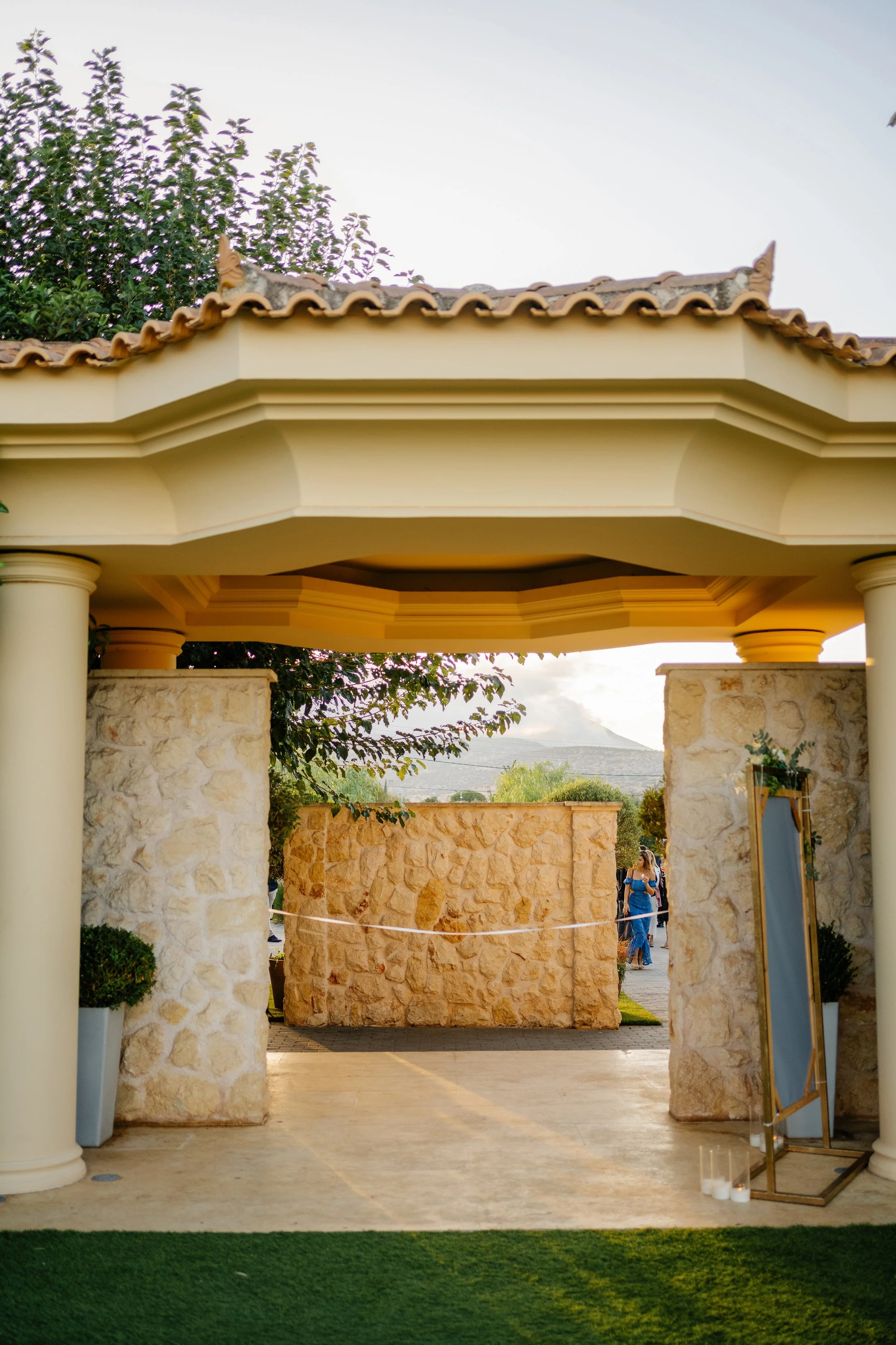 An outdoor wedding venue featuring stone pillars, a beige pergola with decorative molding, and a stone wall with a backdrop of trees and mountains. Some guests are visible in the background.
