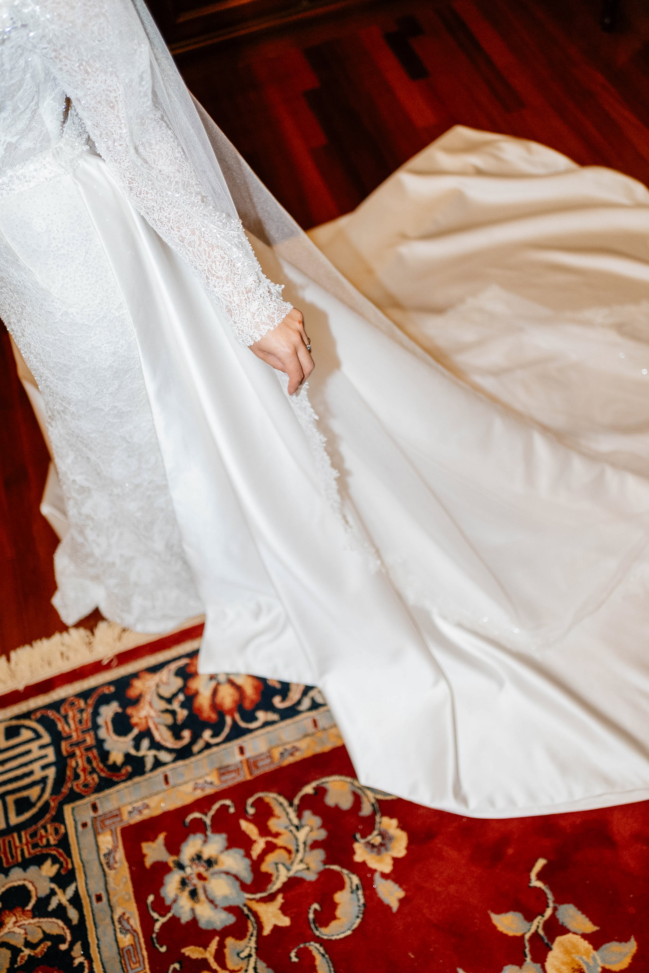 A white wedding gown with lace details and a satin ribbon, partially on an ornate red and blue patterned rug.