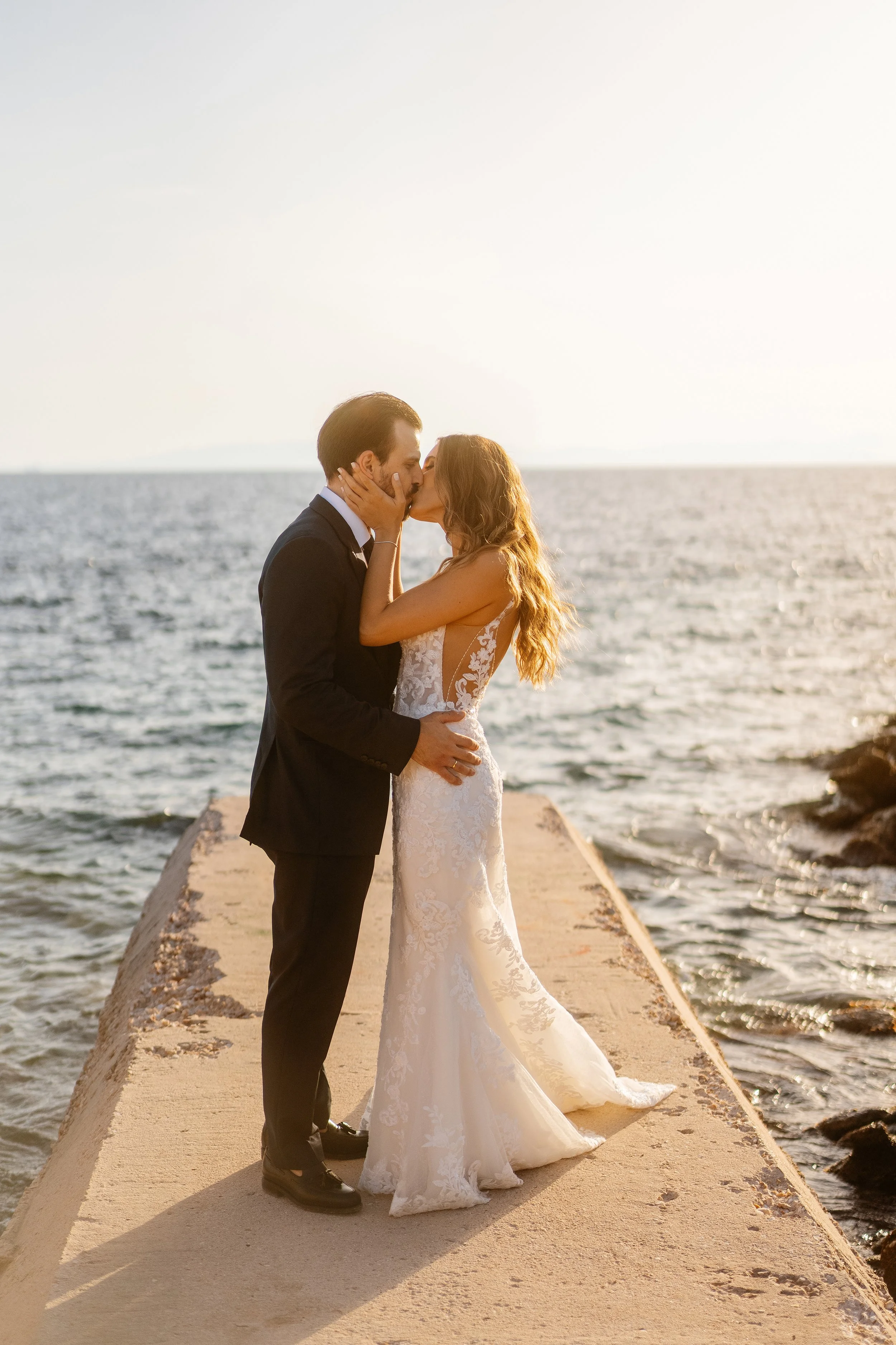 A bride and groom sharing a romantic kiss on a pier by the ocean at sunset, with the bride in a white lace gown and the groom in a black tuxedo.