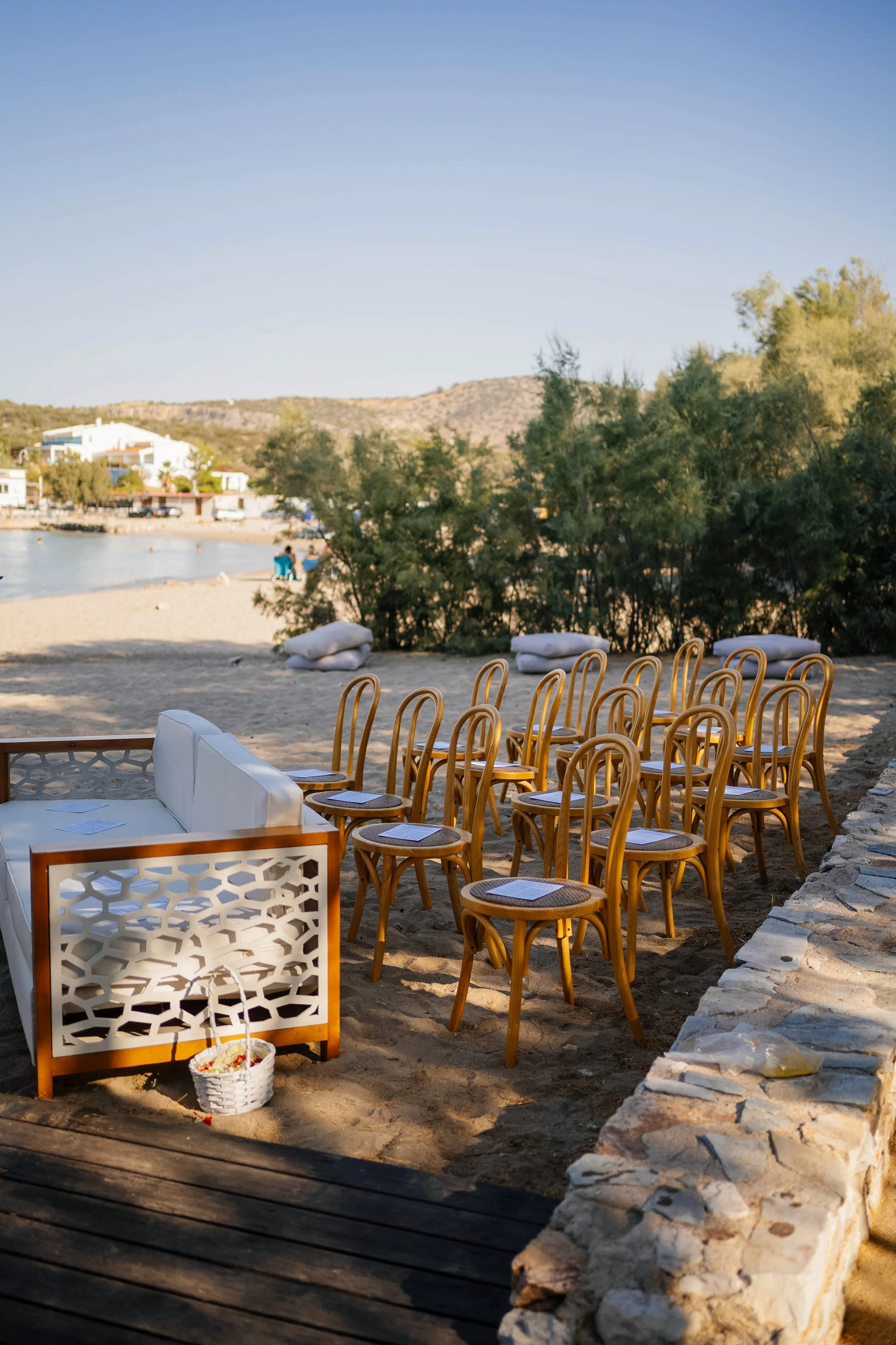 Empty outdoor seating area with wooden chairs and white cushions on a sandy beach, with a basket of flowers in the foreground and a view of water, trees, and hills in the background.