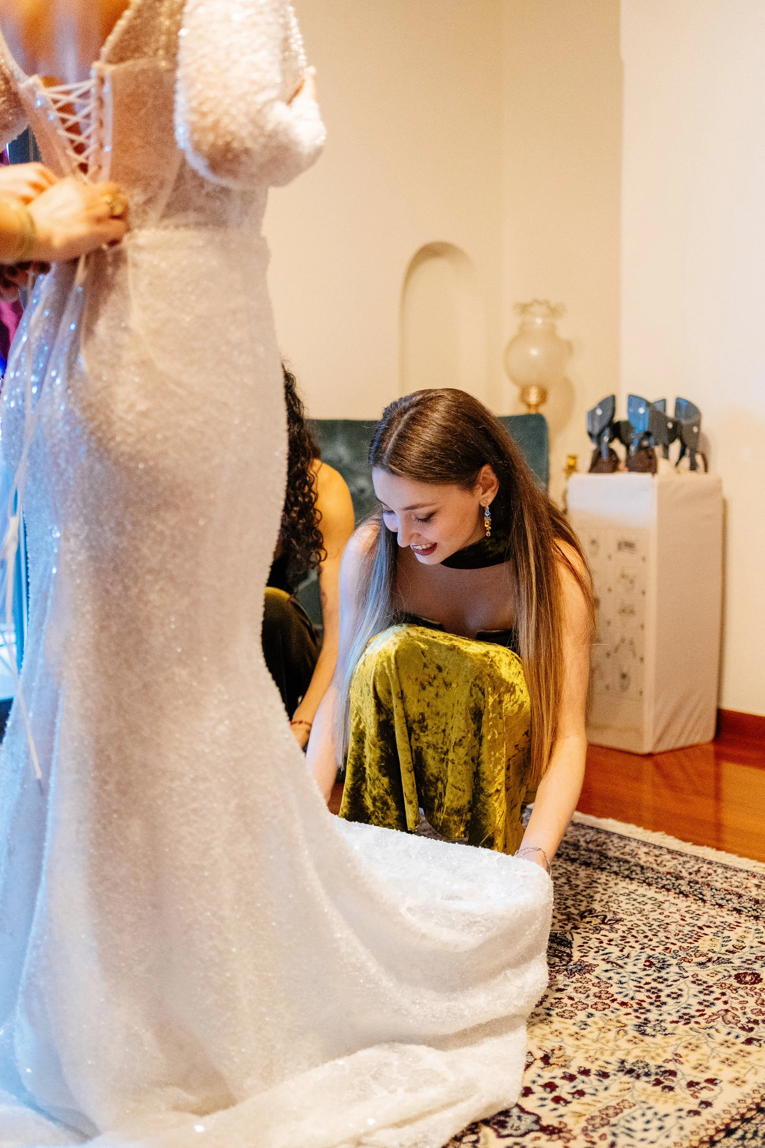 A woman with long brown hair wearing earrings and a sleeveless top looking down and smiling, surrounded by fabric and objects in a room decorated with a patterned rug, a white cabinet with figurines, and a lamp with a glass base.