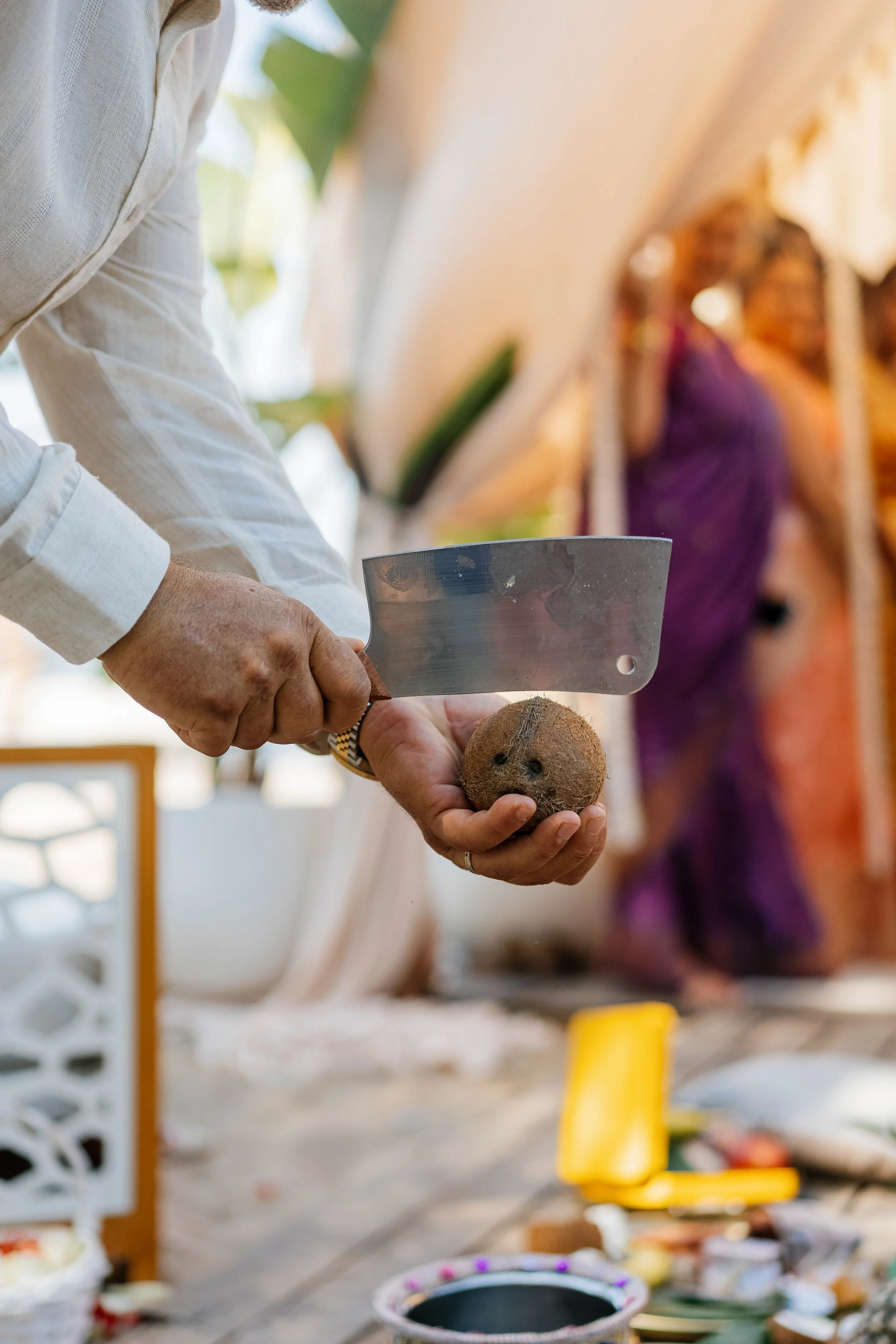 Person holding a coconut and knife, preparing to cut it, with people and colorful objects in the background.