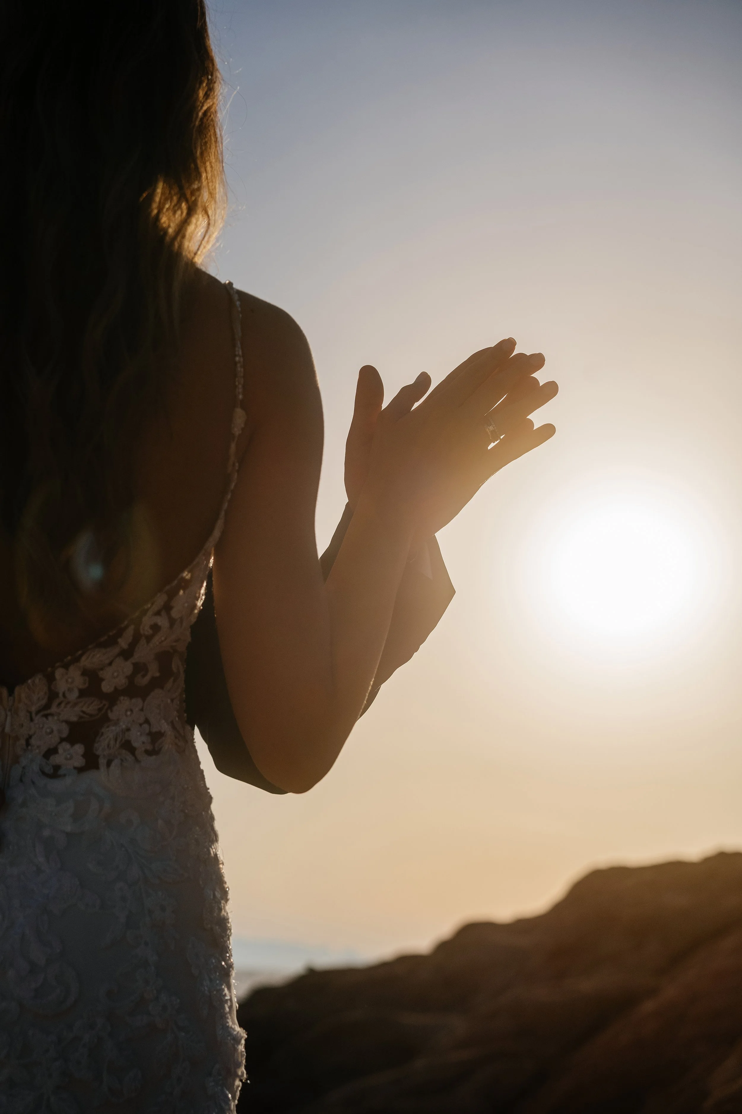 A woman with long hair, dressed in a lace gown, is praying with her hands folded in front of her during sunset or sunrise by the mountains.