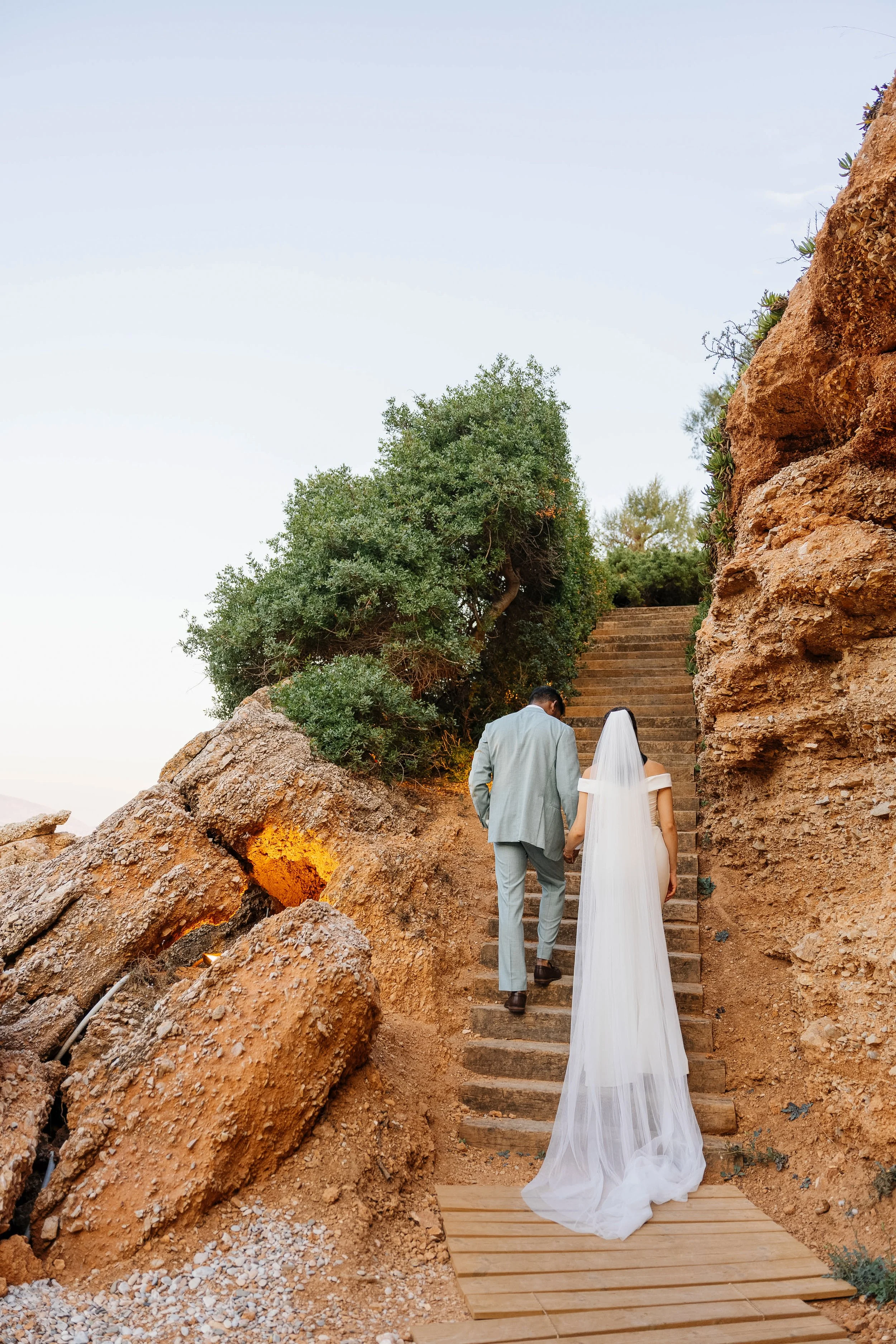 A bride and groom holding hands, walking up stone stairs outdoors between rocky formations and green trees, during sunset or late afternoon.