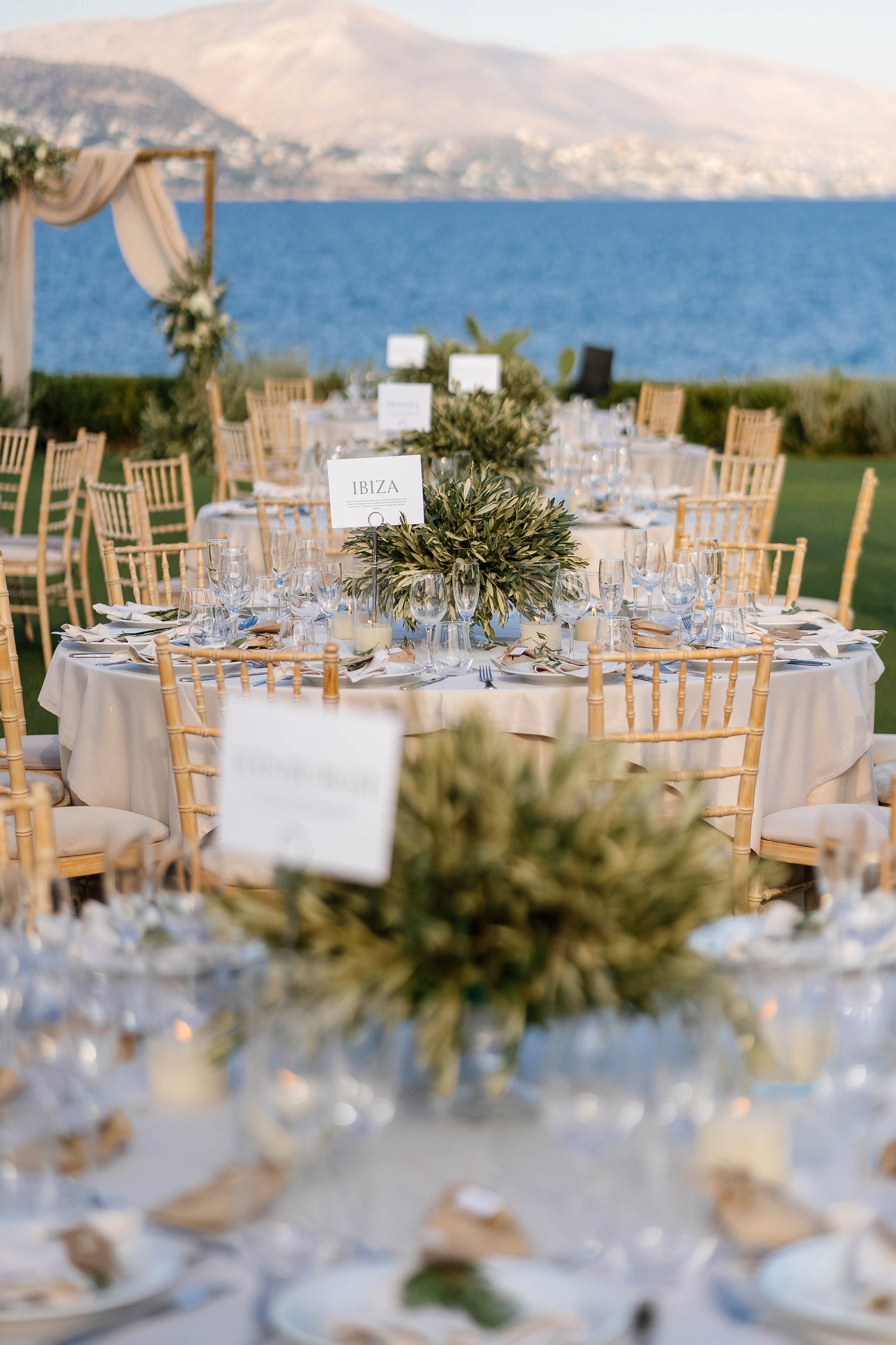 Outdoor wedding reception setup on a lawn overlooking a large body of water with mountains in the background. Round tables with white tablecloths, glassware, and greenery centerpieces, surrounded by wooden chairs, and a wooden arch decorated with fab