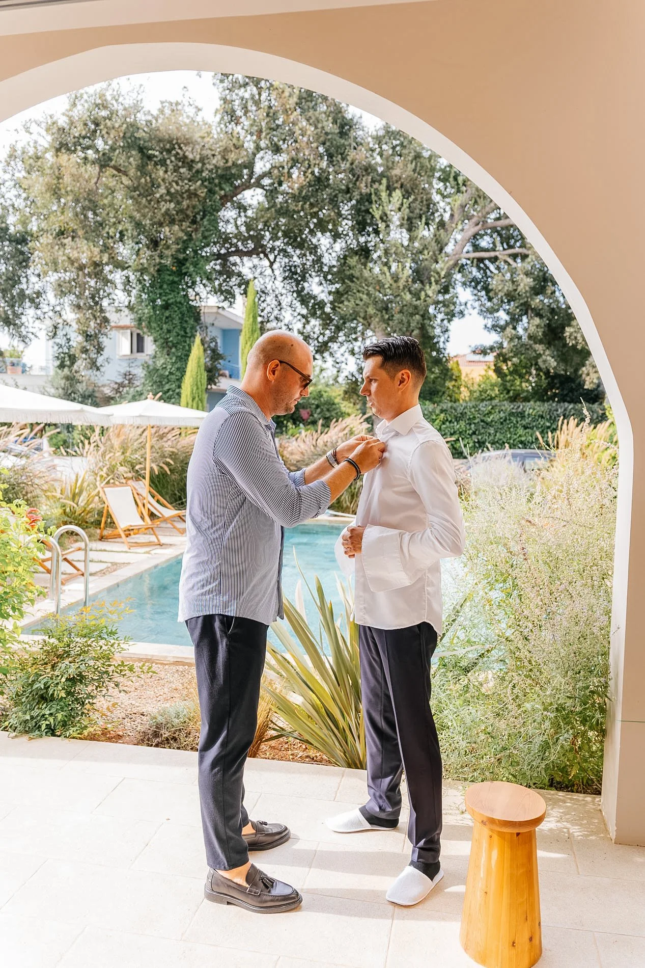 Two men standing in a garden, one helping the other adjust his white dress shirt, near a pool and outdoor seating area, with trees and plants in the background.