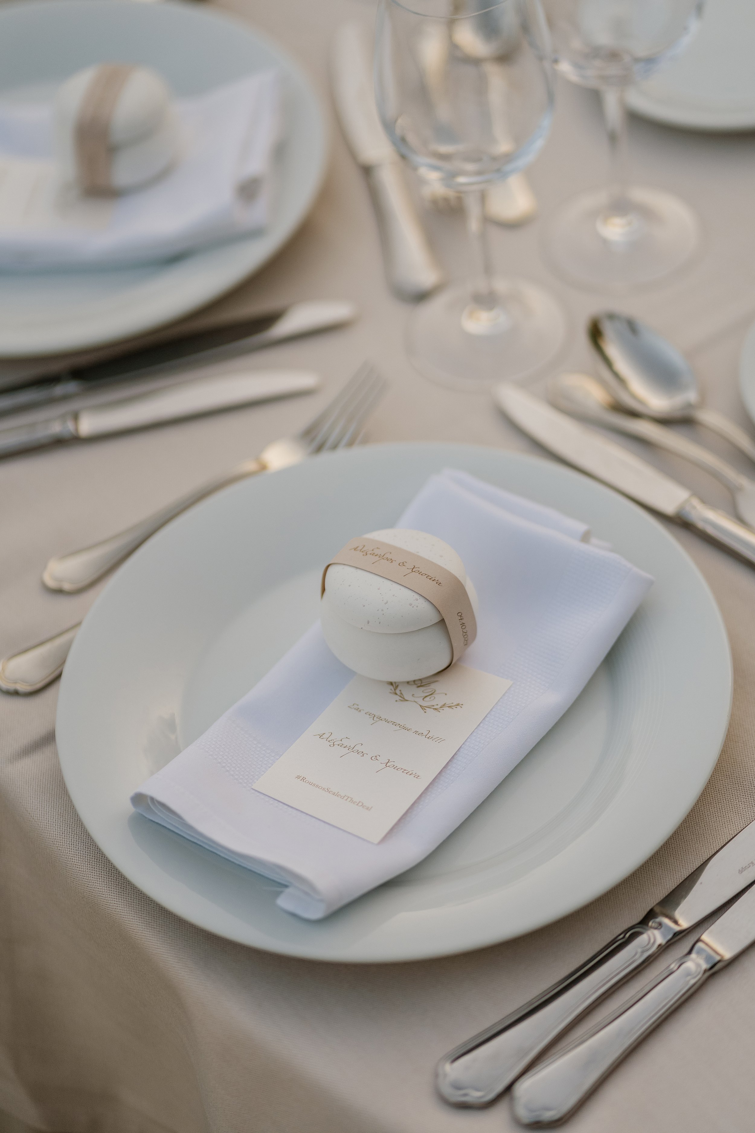 A formal table setting featuring a white plate, a folded white napkin with a printed menu or note, silverware including a knife, fork, and spoon, and a set of empty wine glasses. The tablecloth is beige.
