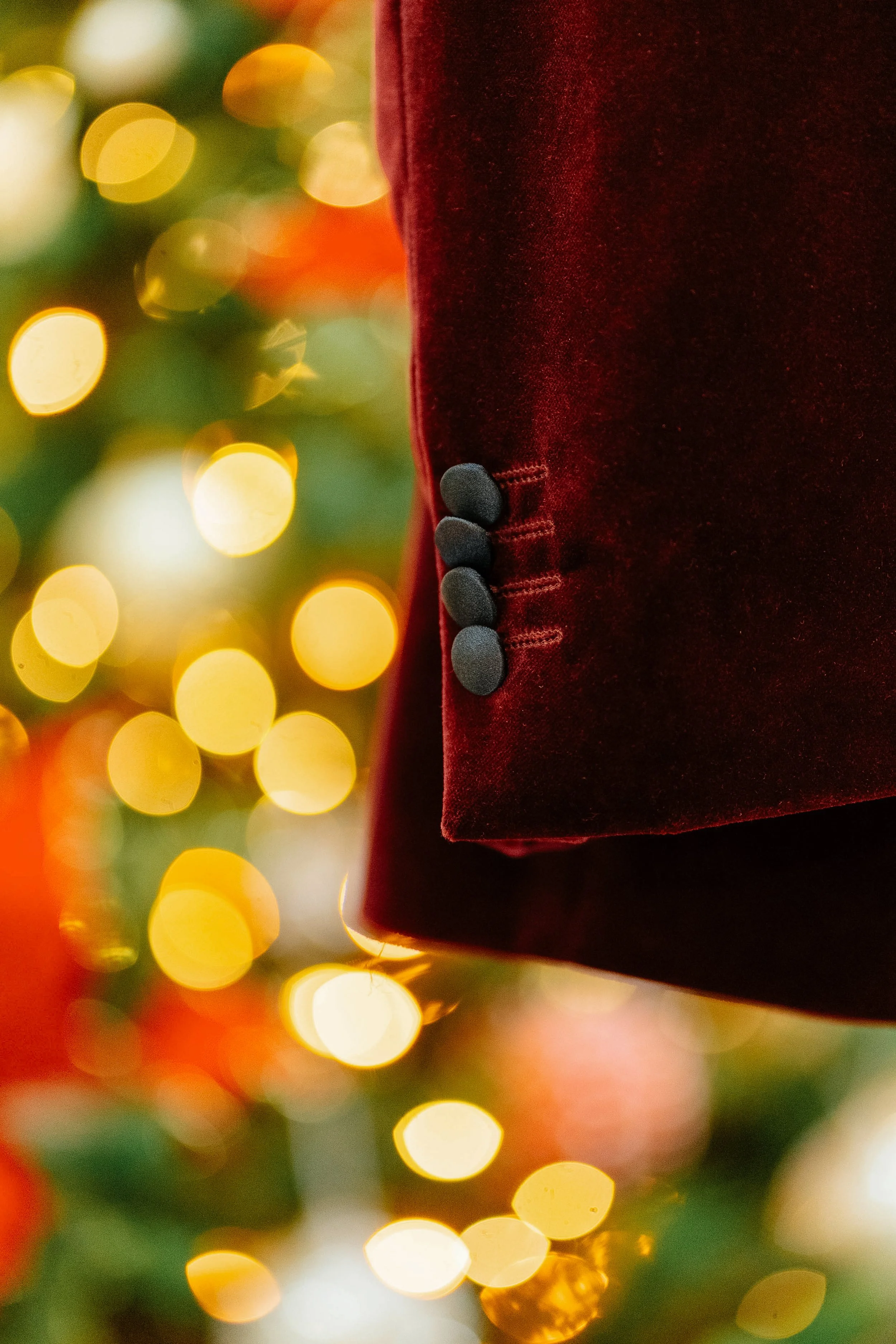 Close-up of a red velvet Christmas ornament with black buttons along the seam, and blurred yellow and green holiday lights in the background.