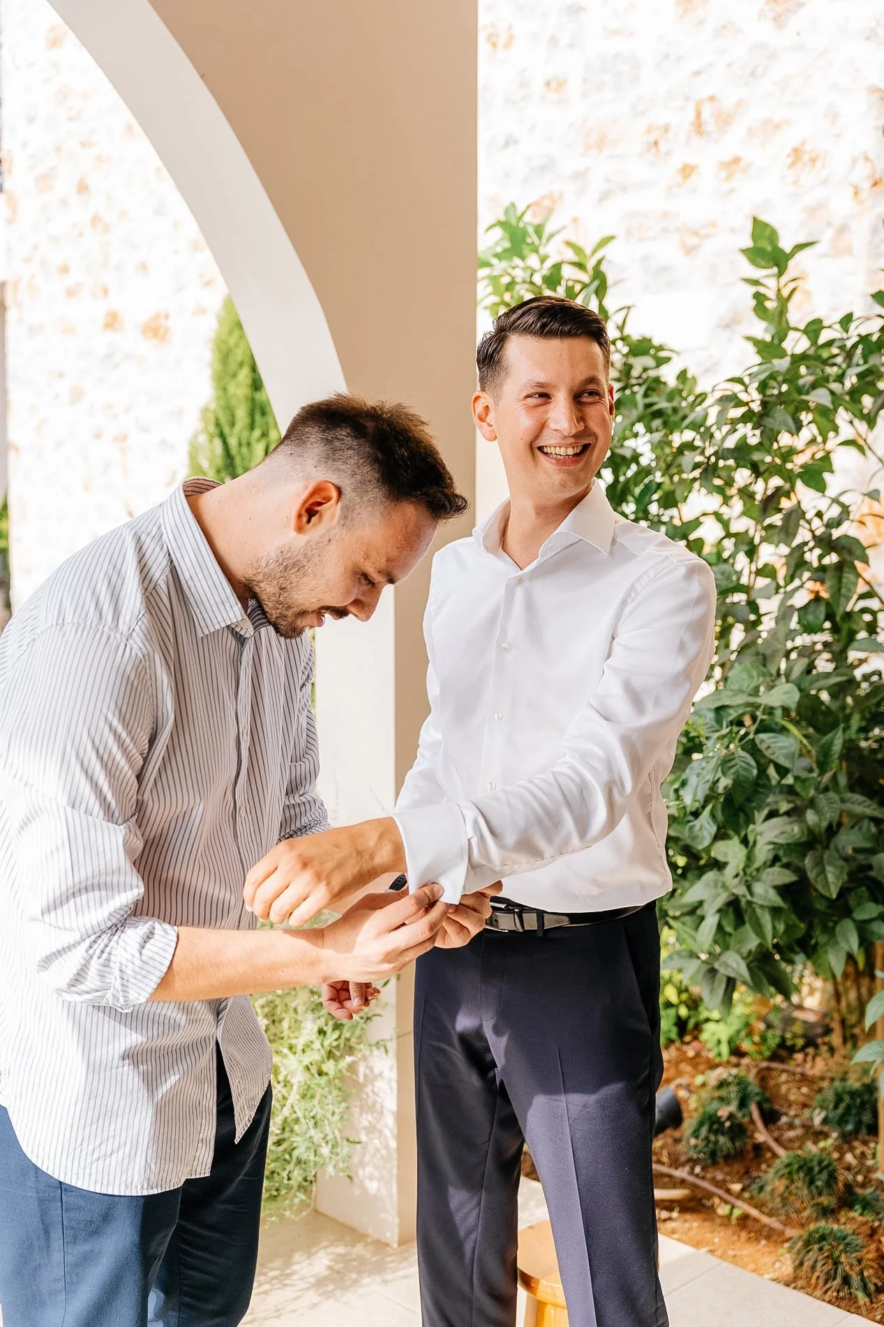 Two men standing outside, one is smiling and wearing a white shirt, the other is looking down and wearing a striped shirt. They are engaged in a moment of handshake or exchange, with greenery and a brick wall in the background.