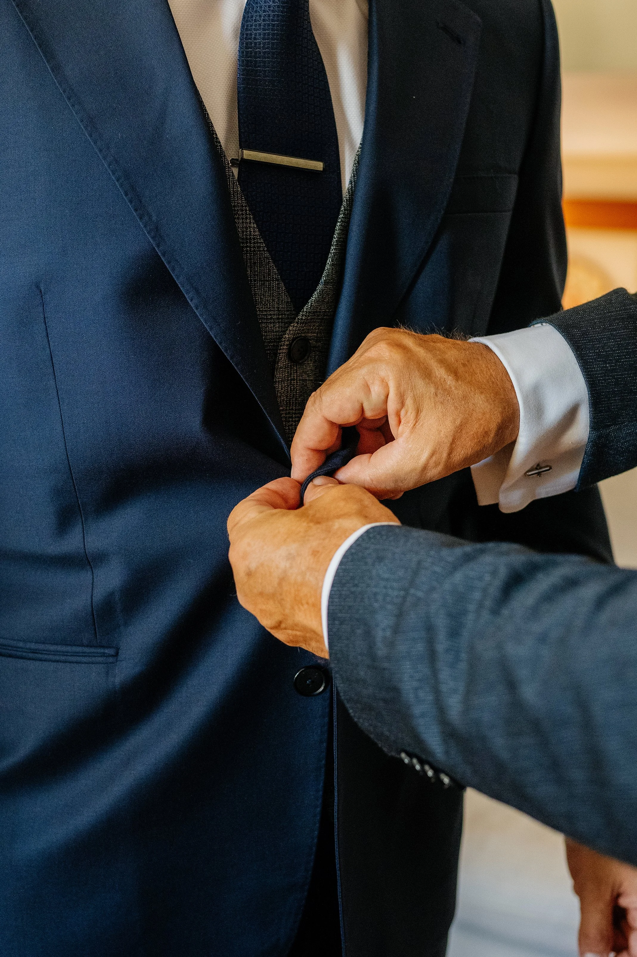 Two men are dressed in formal suits, one in a dark blue suit and the other in a gray suit, with the man in the dark blue suit buttoning it up while the man in gray helps. The man in the dark blue suit is wearing a white shirt, a dark blue tie, and a 