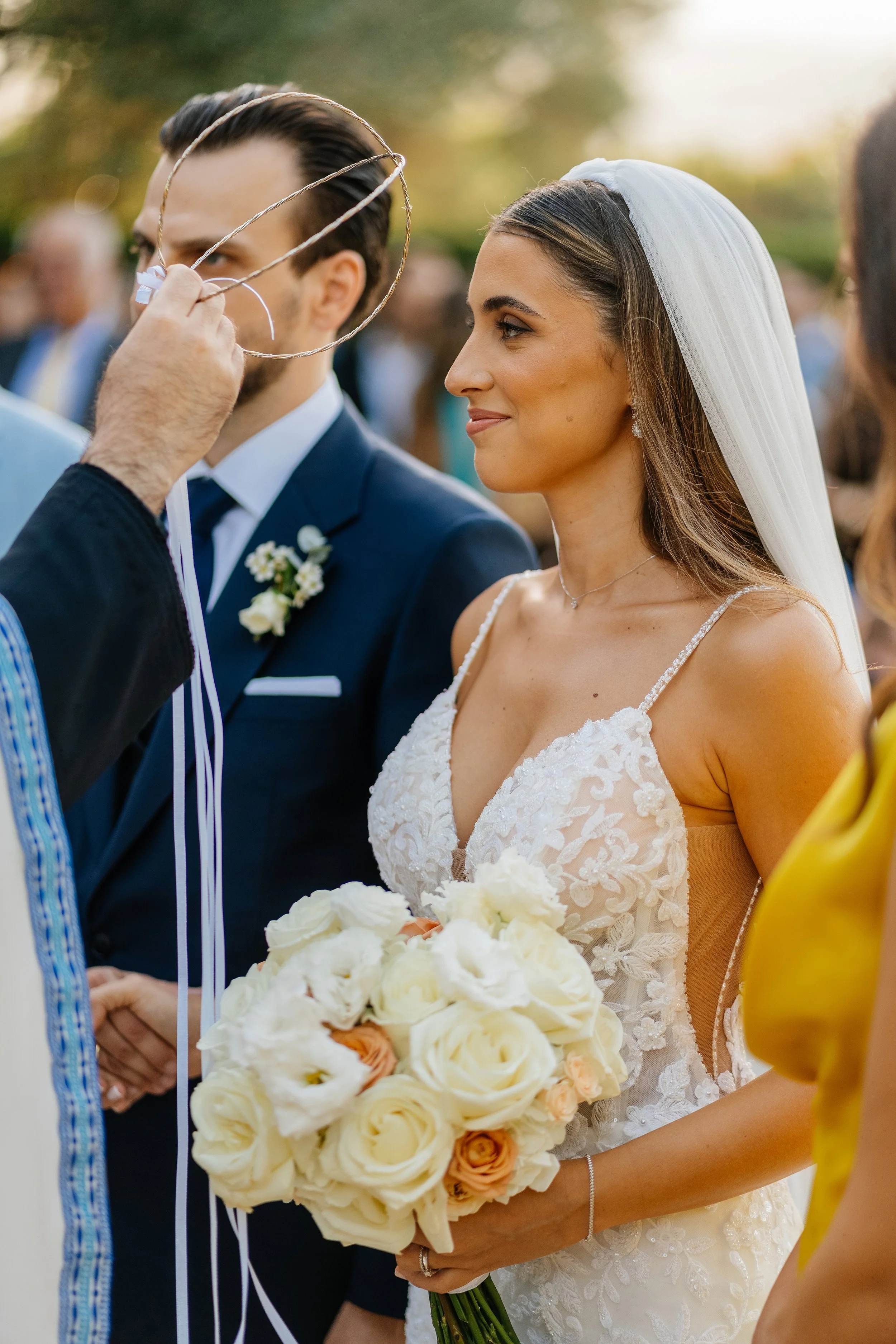 A bride in a white wedding dress holding a bouquet of white and peach roses, smiling as she stands next to a groom during a wedding ceremony.