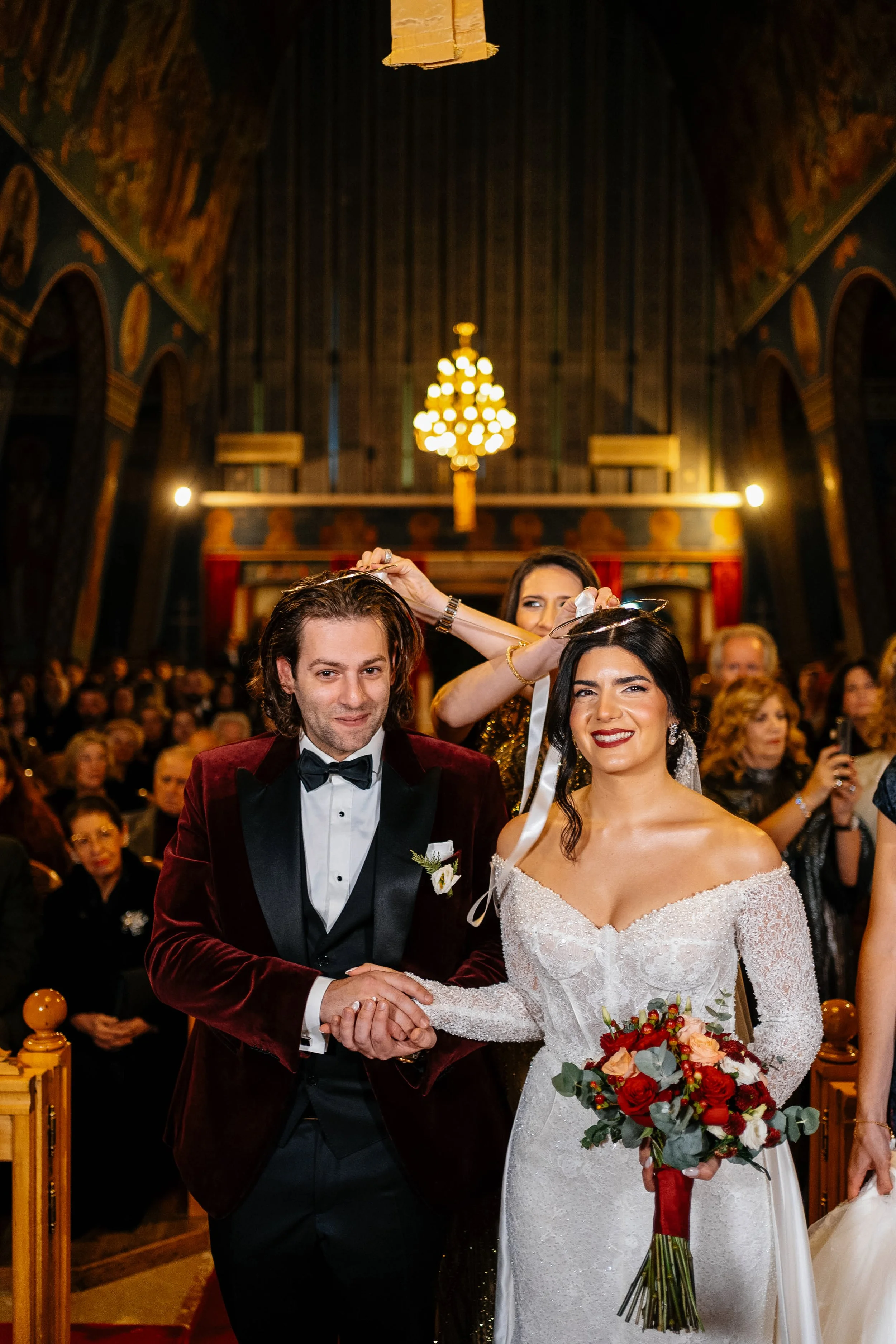 A couple is getting married in a church, holding hands and facing the camera. The groom is wearing a dark red velvet tuxedo jacket with a black bow tie, and the bride is holding a bouquet of red, white, and pink flowers. There are two women behind th
