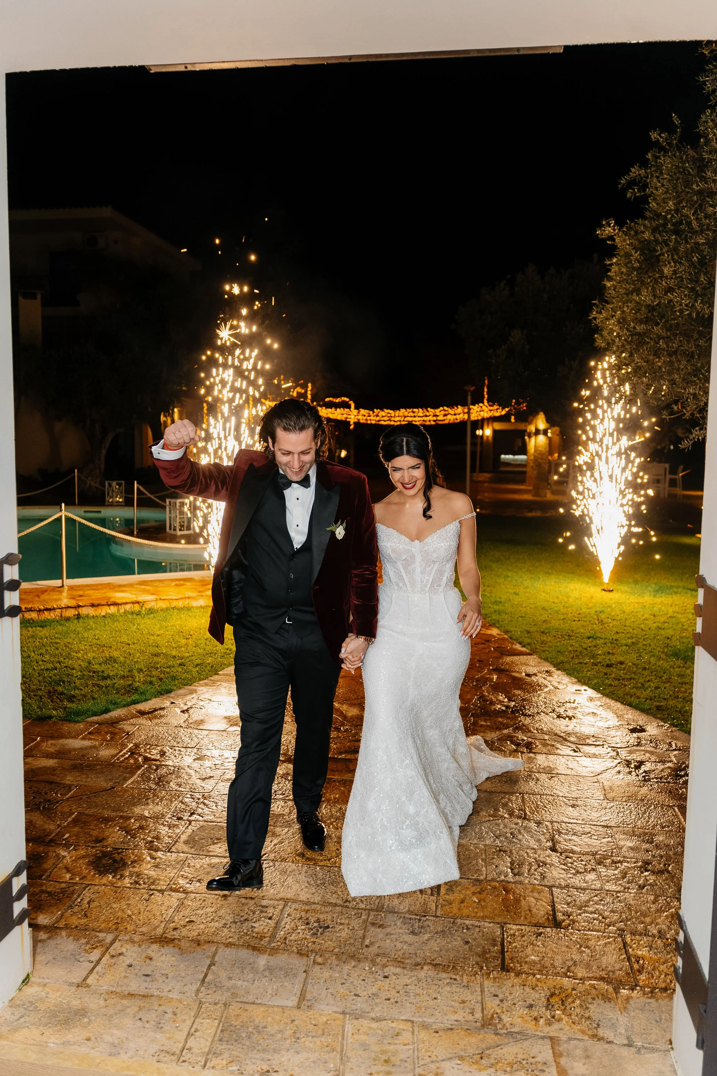 A newlywed couple walking hand-in-hand outdoors at night, celebrating with fireworks and sparklers in the background.