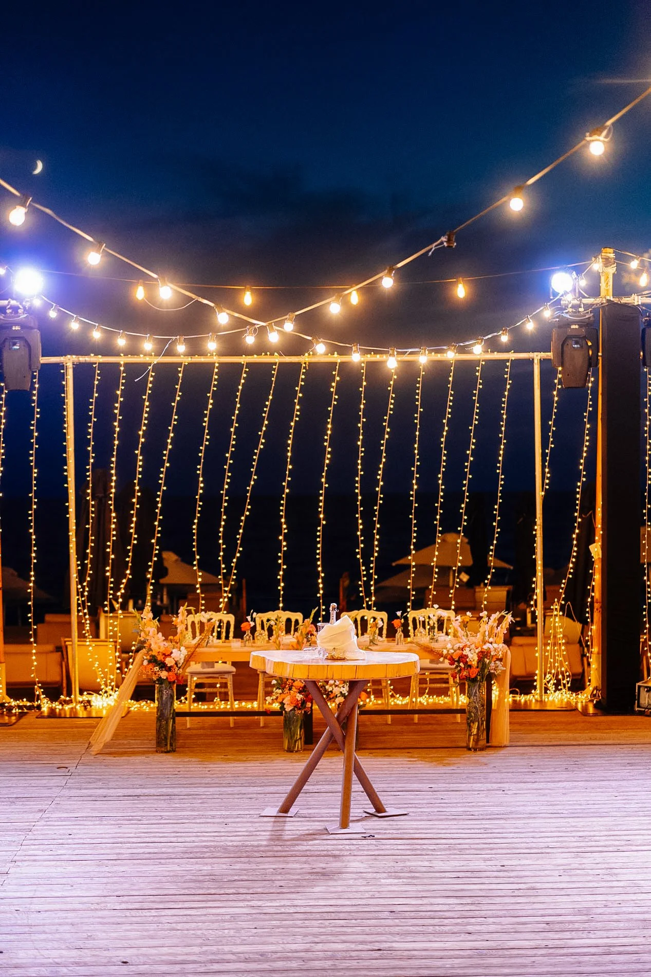 String lights hanging above a decorated outdoor dining area at night, with a table and chairs, floral arrangements, and a nighttime sky.