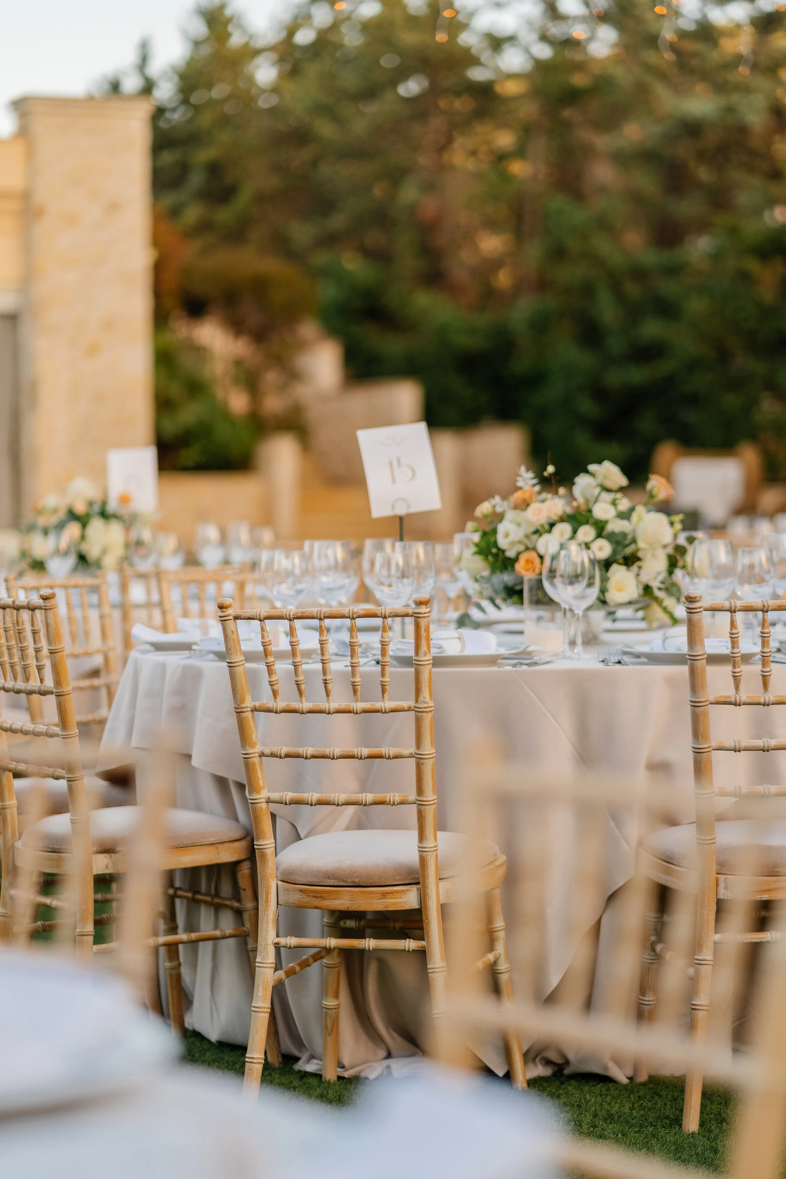 An outdoor event setup with round tables covered in white tablecloths, decorated with floral centerpieces featuring white and peach-colored flowers, surrounded by wooden chairs. One table has a table sign with the number 15. The background shows tree