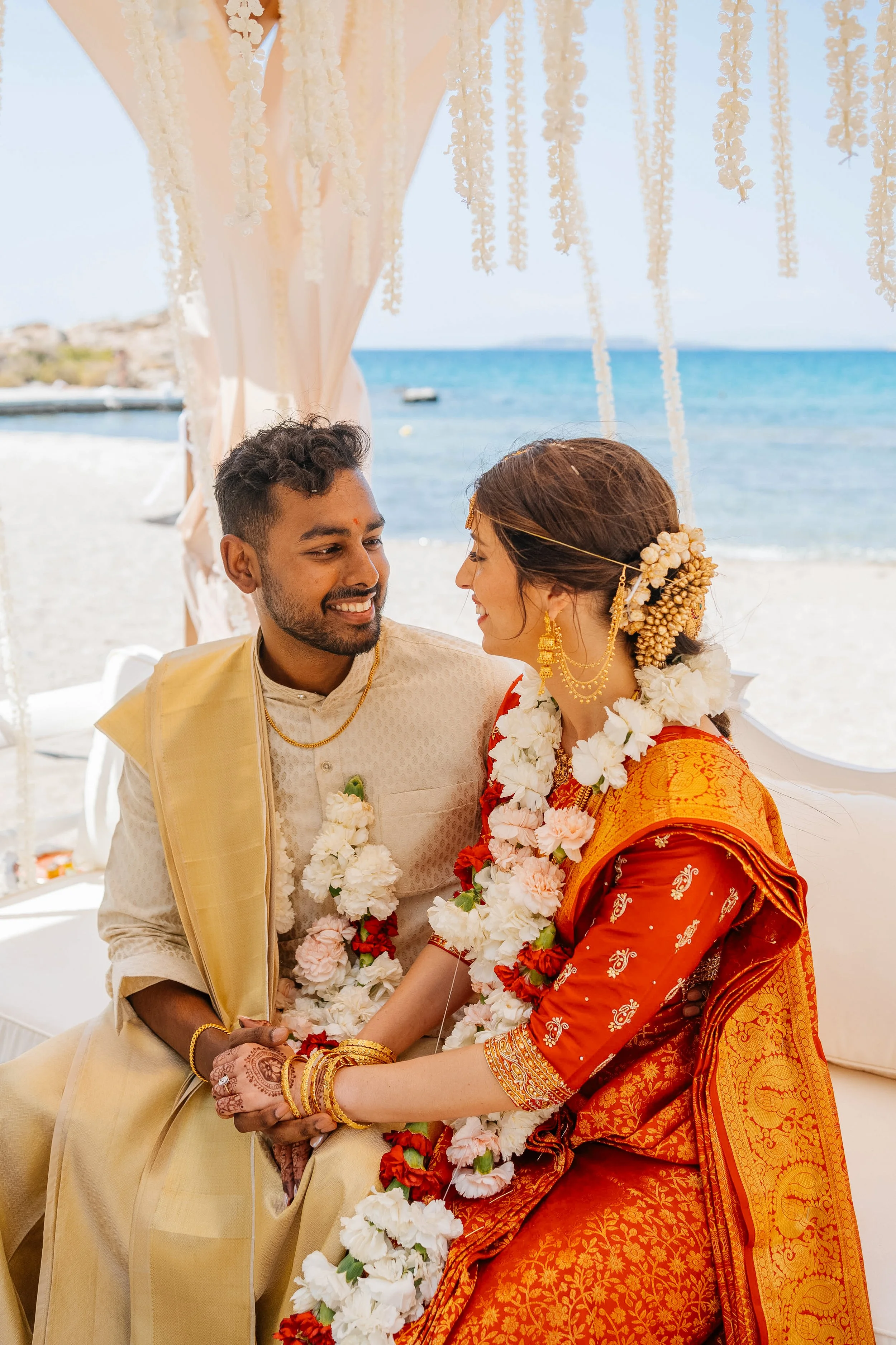 A couple dressed in traditional Indian wedding attire sitting on a beach, holding hands, with a backdrop of the ocean and a decorated canopy.