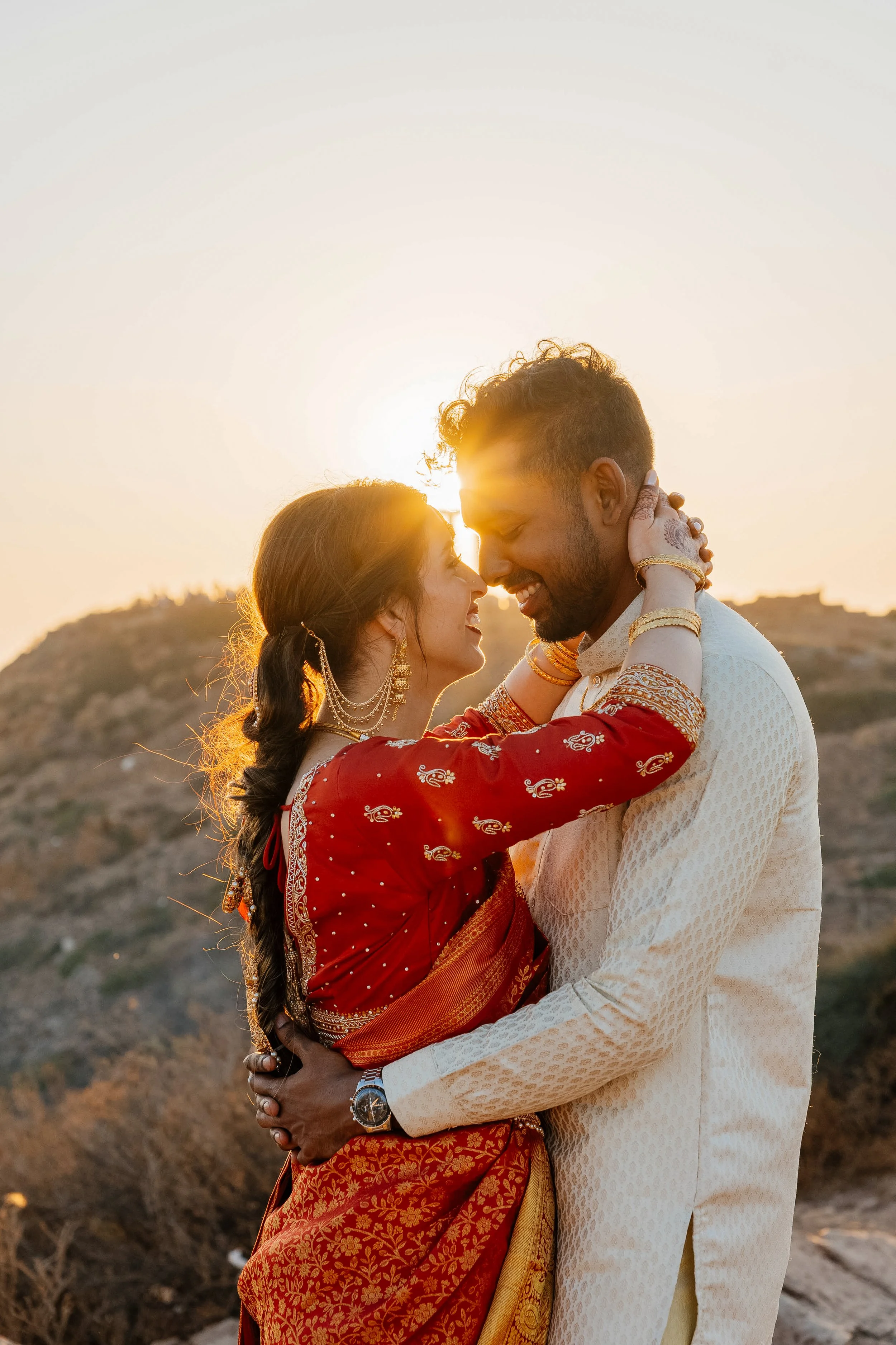 A couple embraces during sunset in a desert-like landscape. The woman wears traditional red and gold Indian attire with jewelry, and the man is dressed in a cream-colored traditional outfit. They are smiling with eyes closed, holding each other close