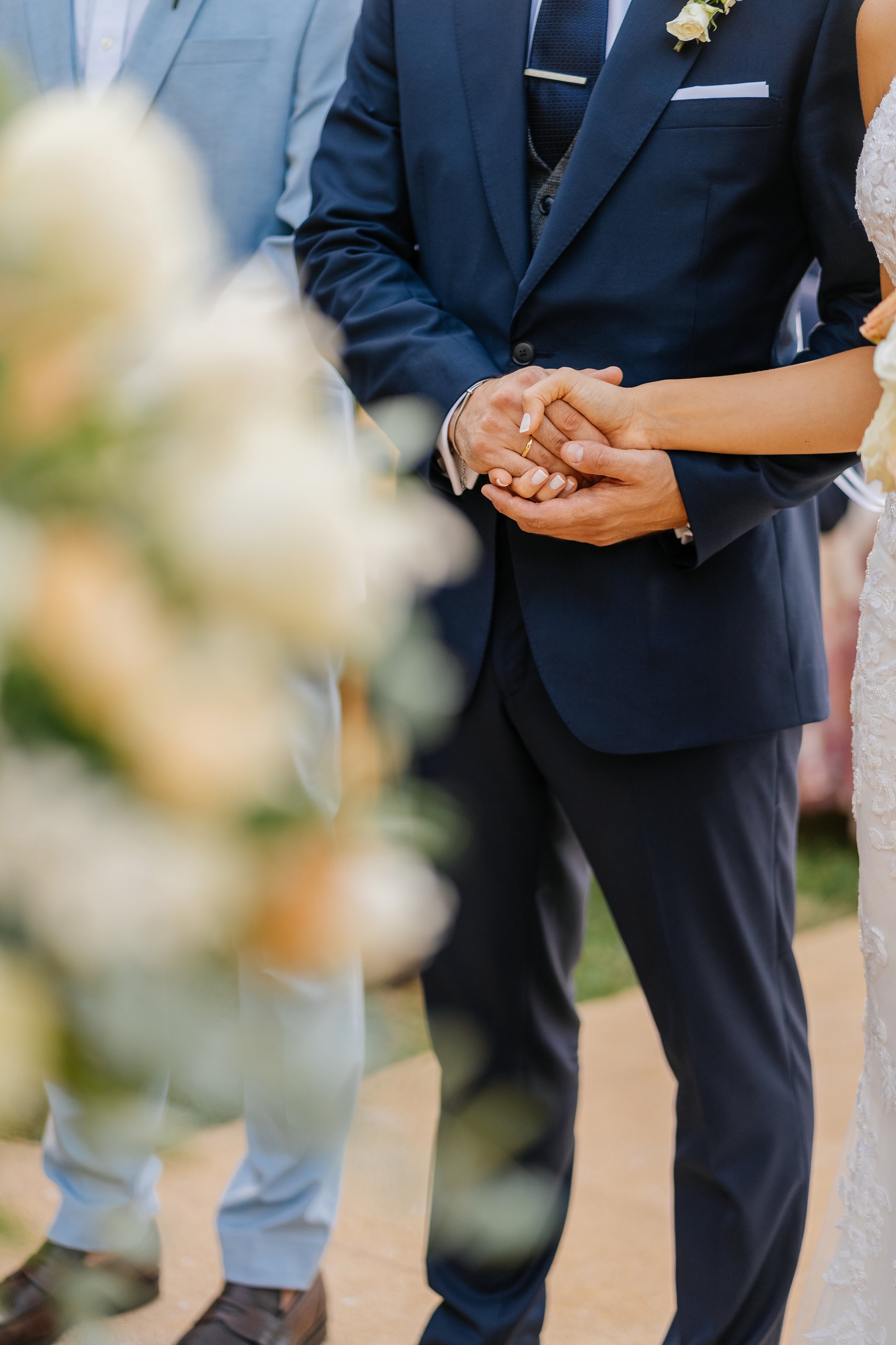 A couple holding hands during a wedding ceremony, dressed in formal attire with a woman in a lace wedding dress and a man in a navy suit with a boutonniere.