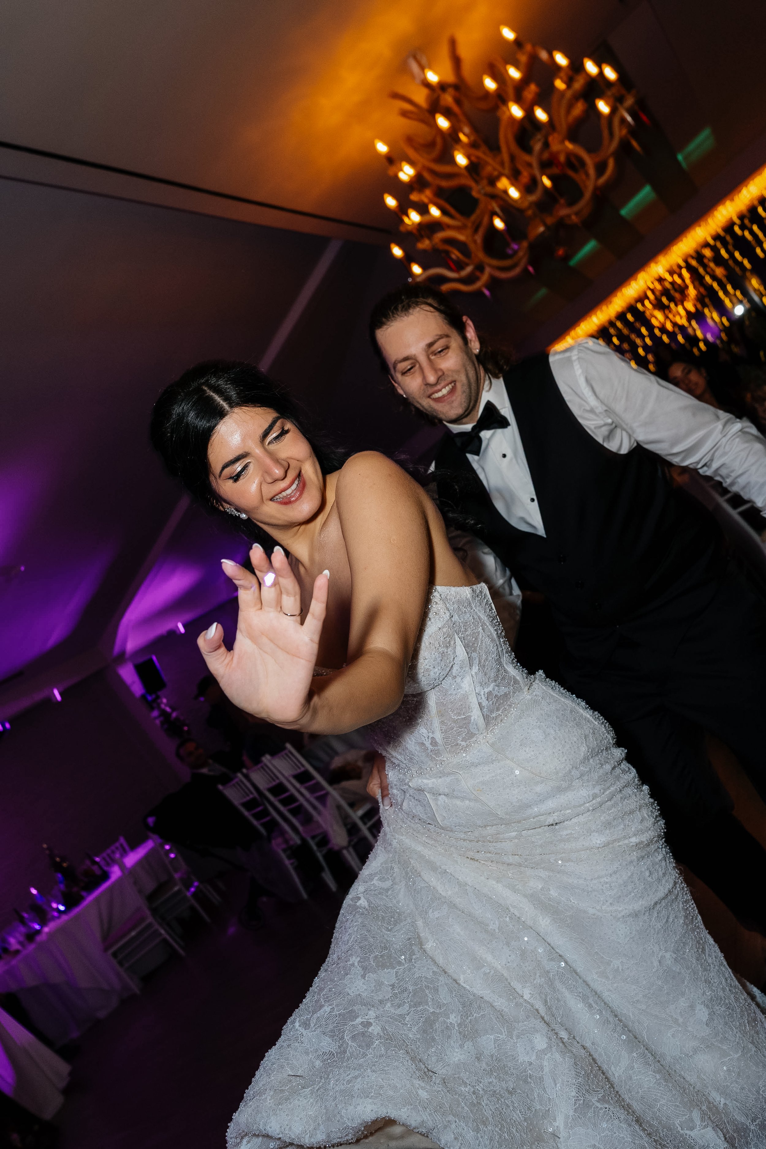 A bride and groom dancing at a wedding reception, with the bride smiling and holding up her hand, and the groom smiling and looking at her, in a decorated banquet hall with chandeliers and purple lighting.