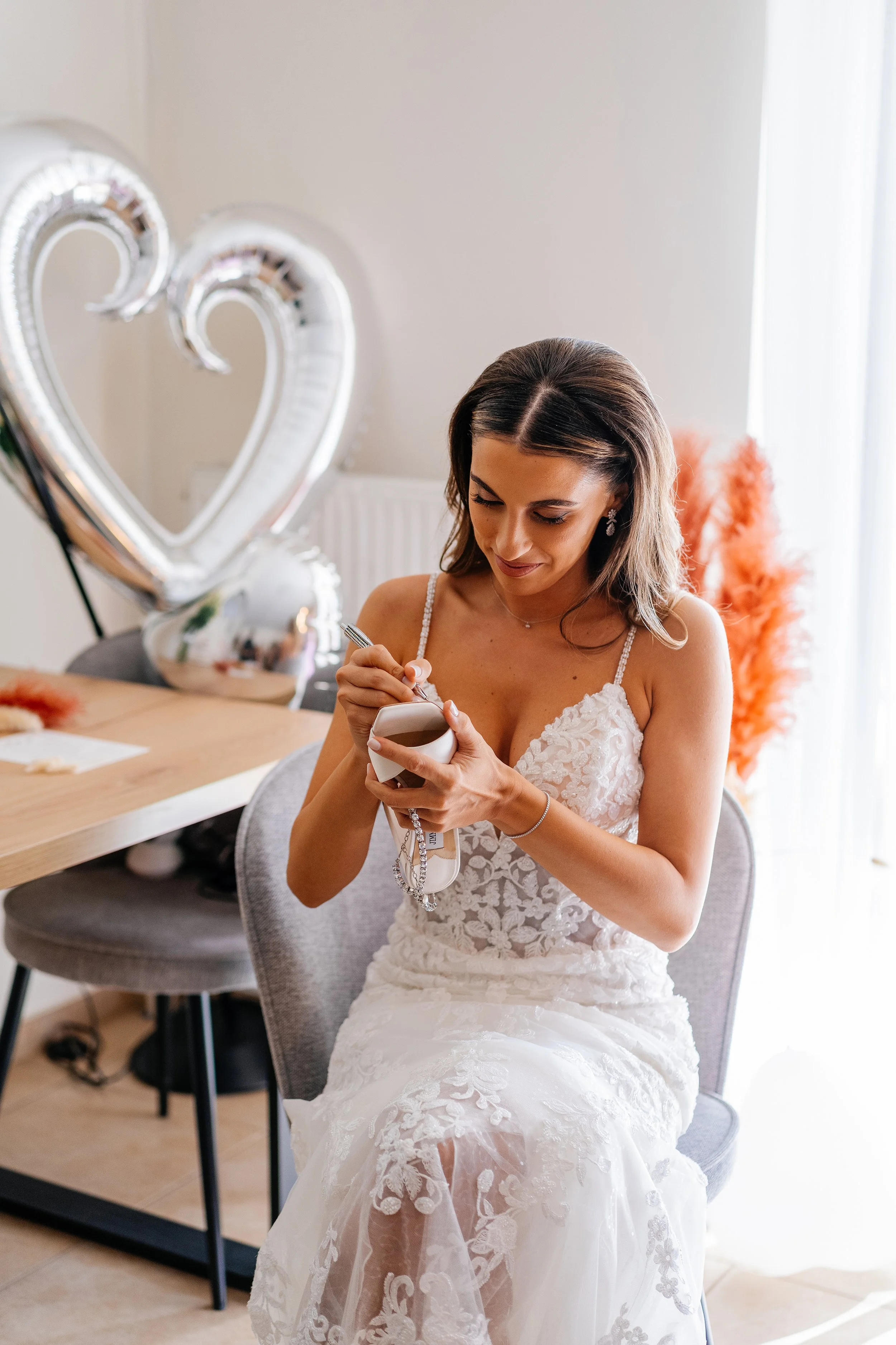 A bride in a white lace wedding dress sitting on a gray chair, holding a phone and a lanyard, with a woman in the background and orange decorative feathers on a stand behind her.