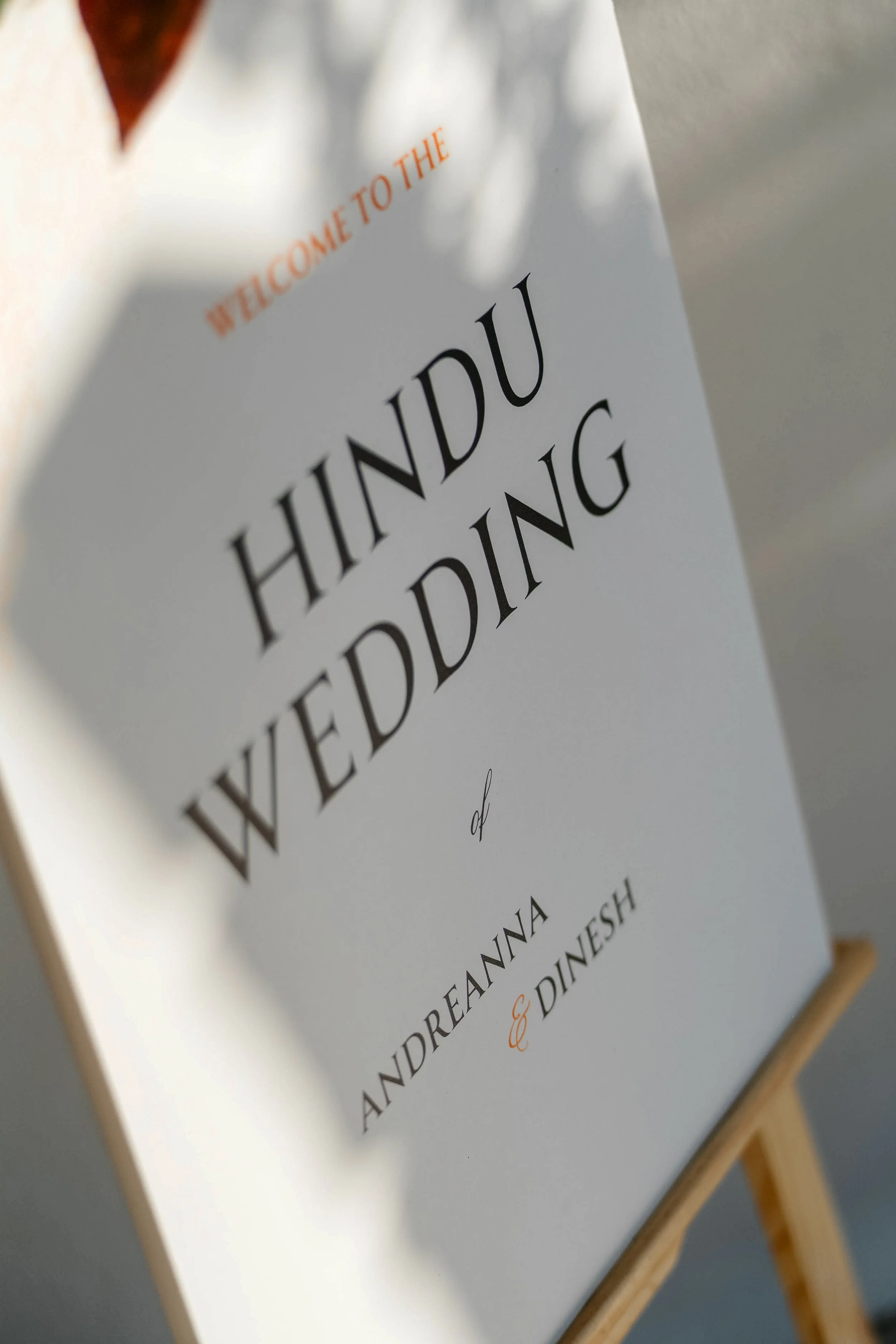 Close-up of a white menu or sign with black and orange text, partially visible, held on a wooden easel. The text reads 'WELCOME TO THE' and 'HINDU EDIDING'.